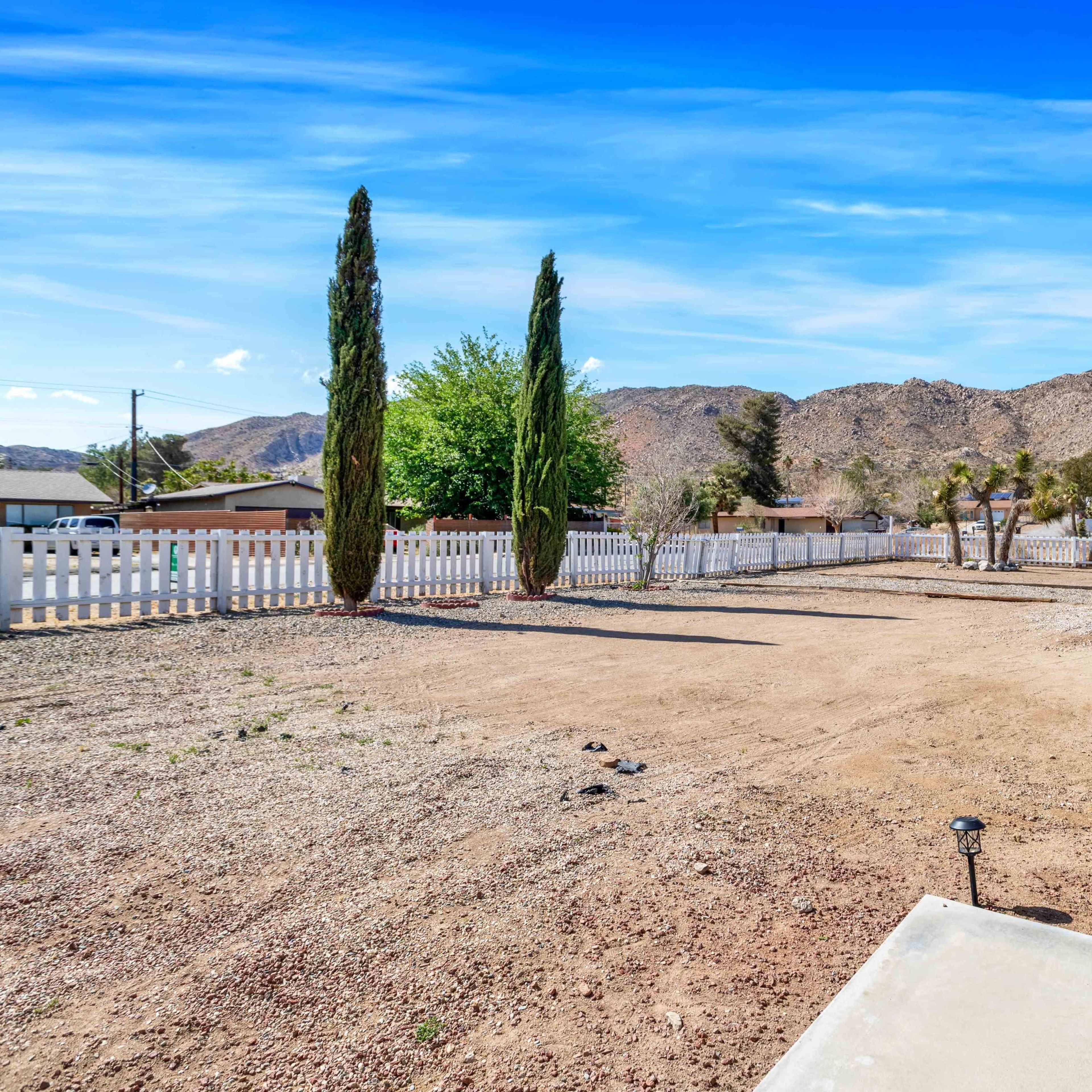An empty, sandy lot is bordered by a white fence and features several trees with mountains in the background.
