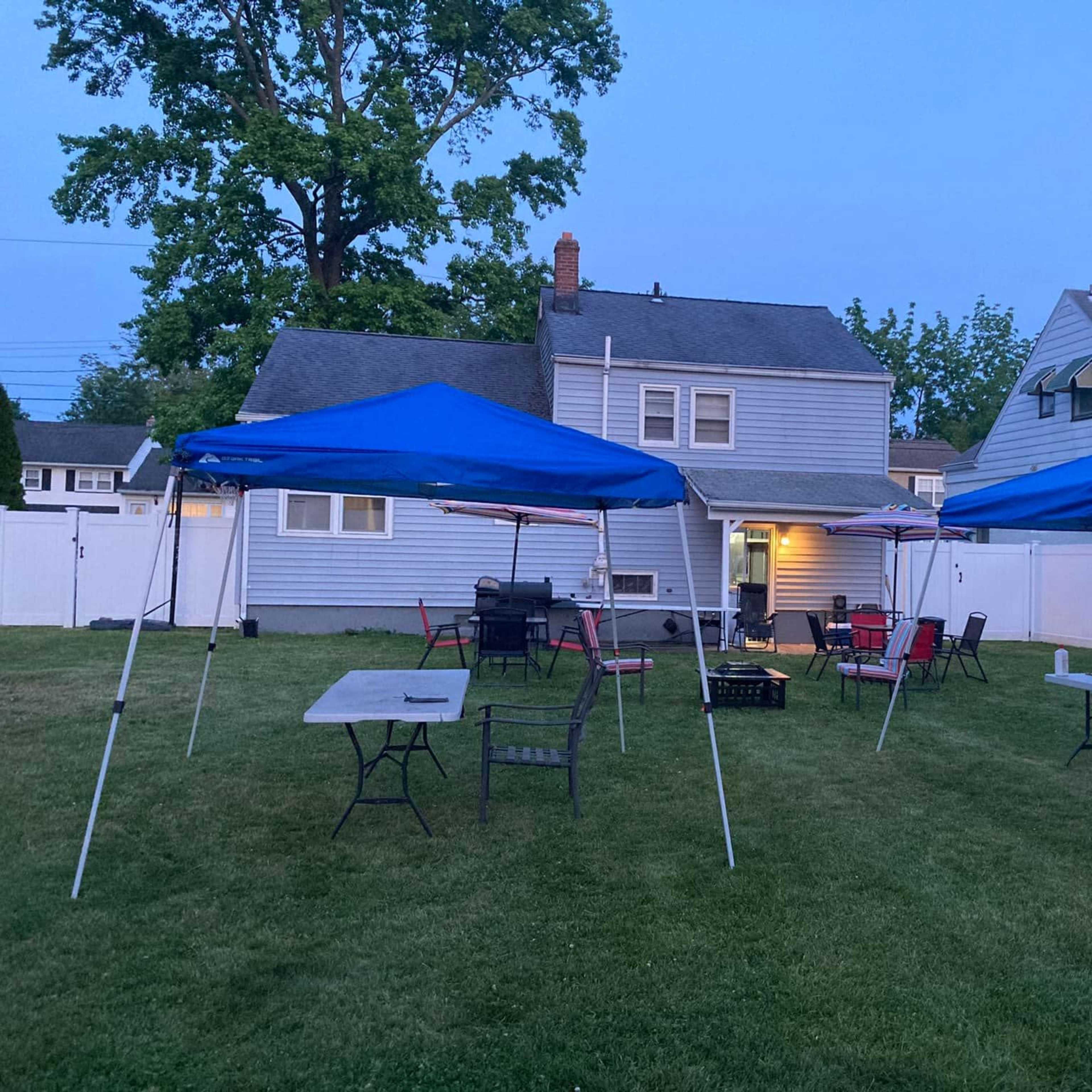 A backyard with two blue party tents set up on a grassy area in front of a house.