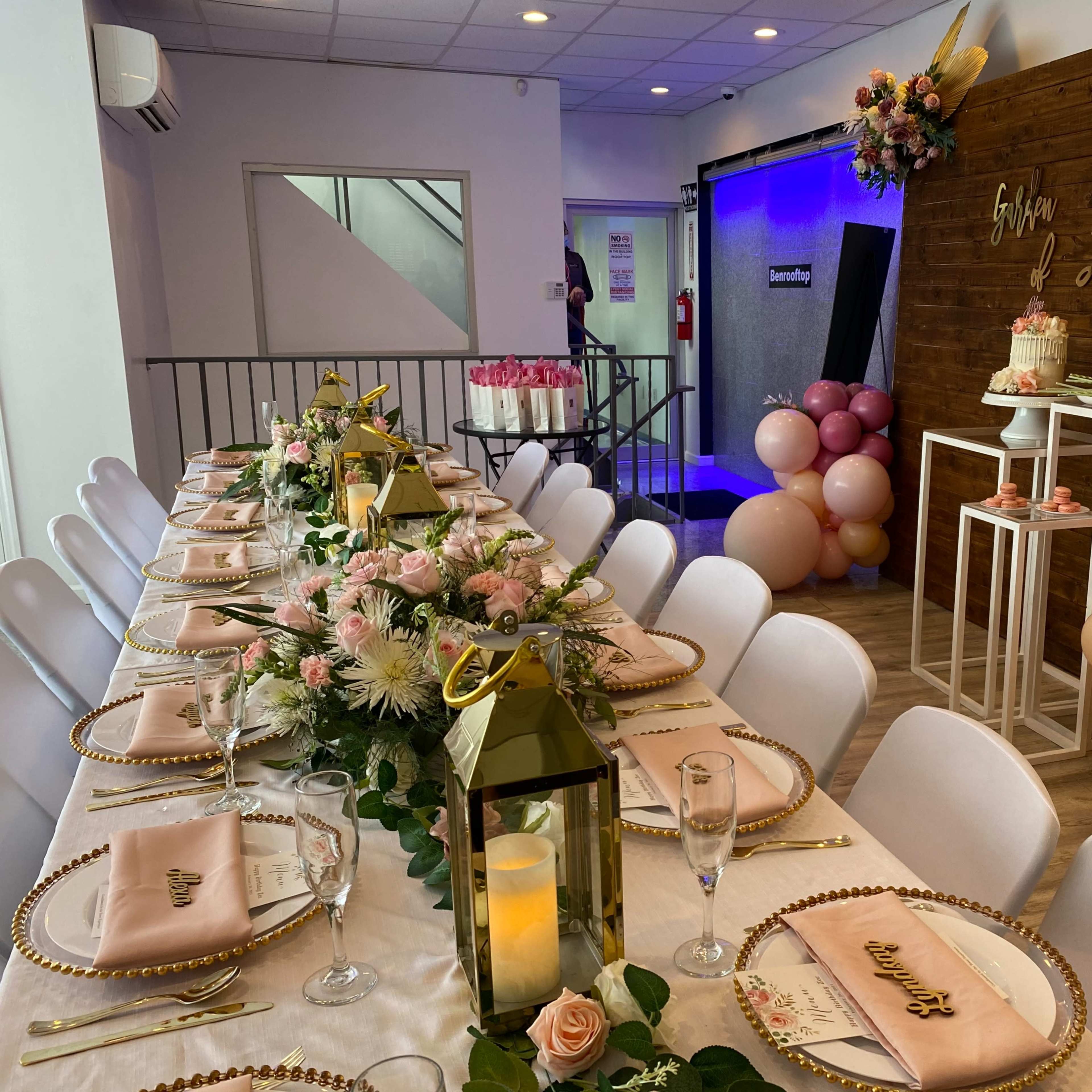 A long dining table is set with decorative tableware and floral arrangements, surrounded by pastel balloons and a dessert display.