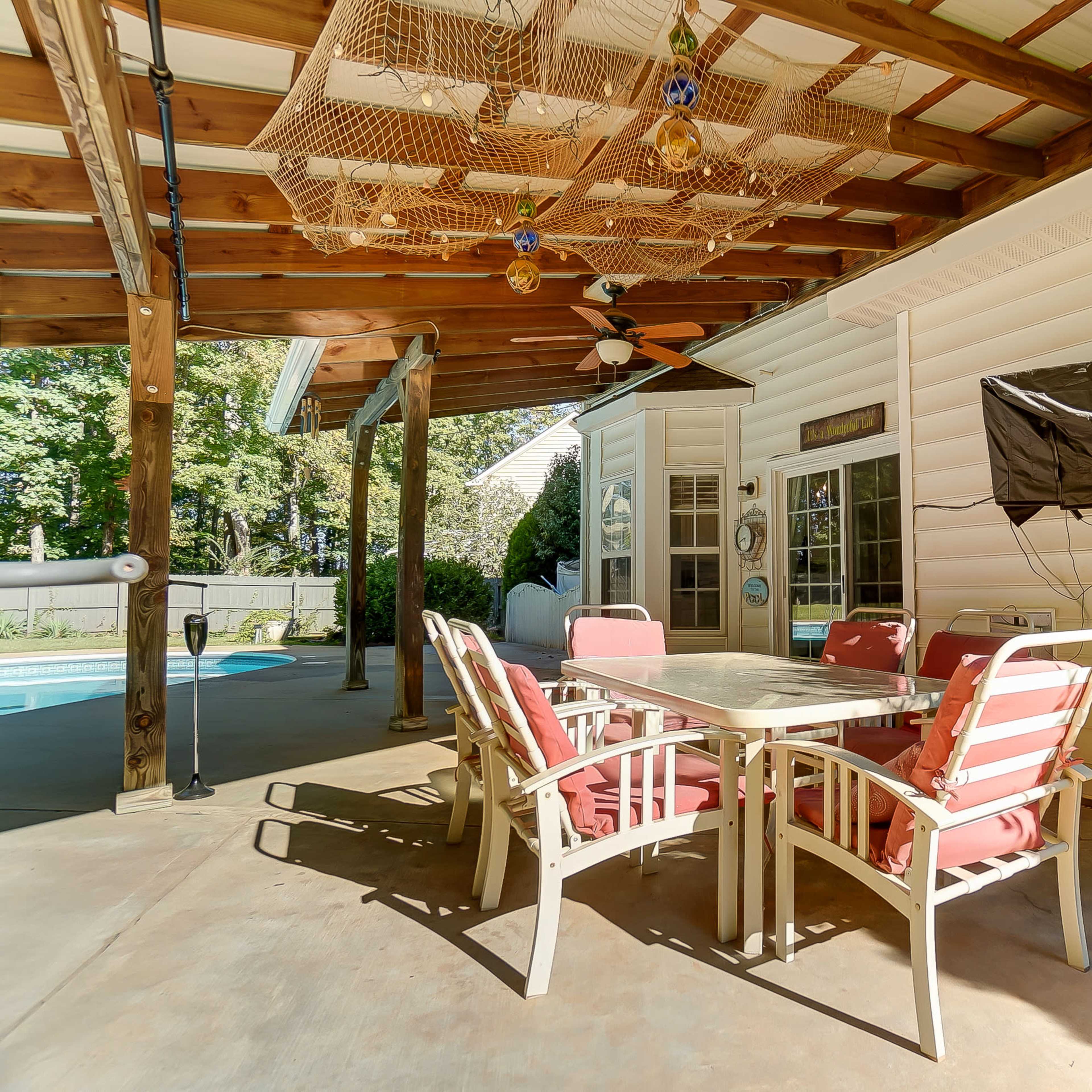 A covered outdoor patio area with a dining table and chairs, a ceiling fan, and a mounted television beside a swimming pool.