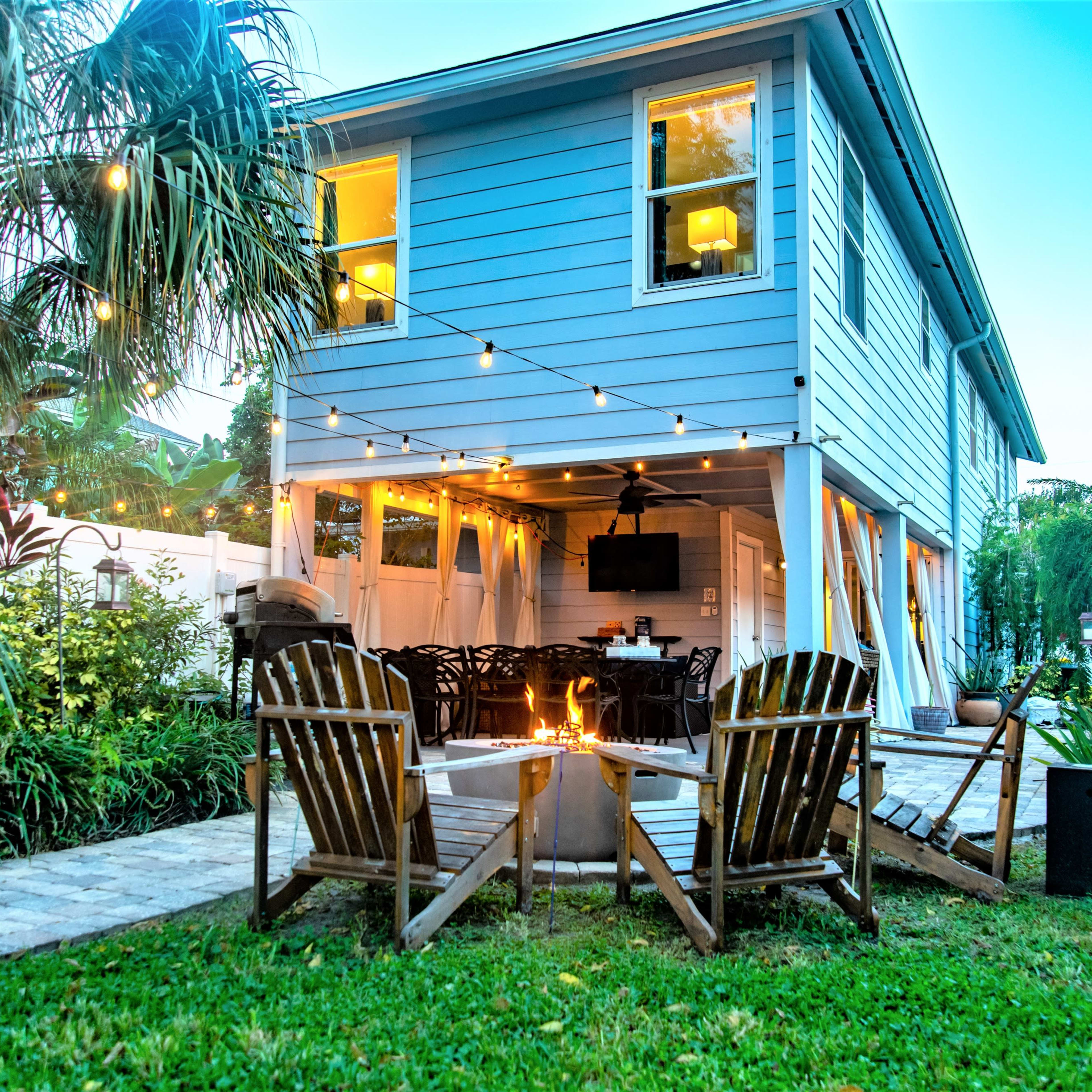 A backyard scene features a blue two-story house with string lights, an outdoor dining area, and a fire pit surrounded by wooden chairs.
