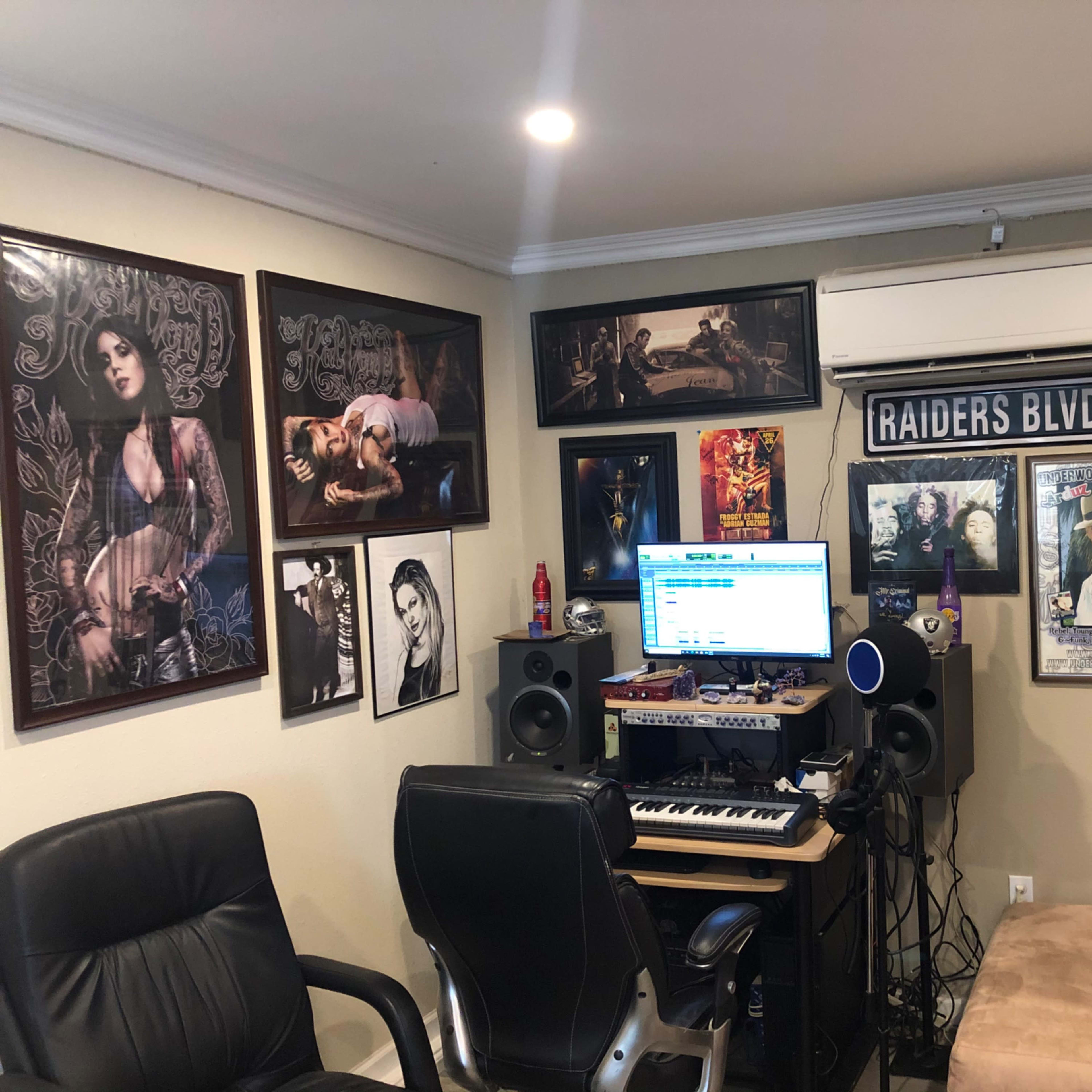 A home recording studio with a computer, keyboards, speakers, and numerous framed posters on the walls.