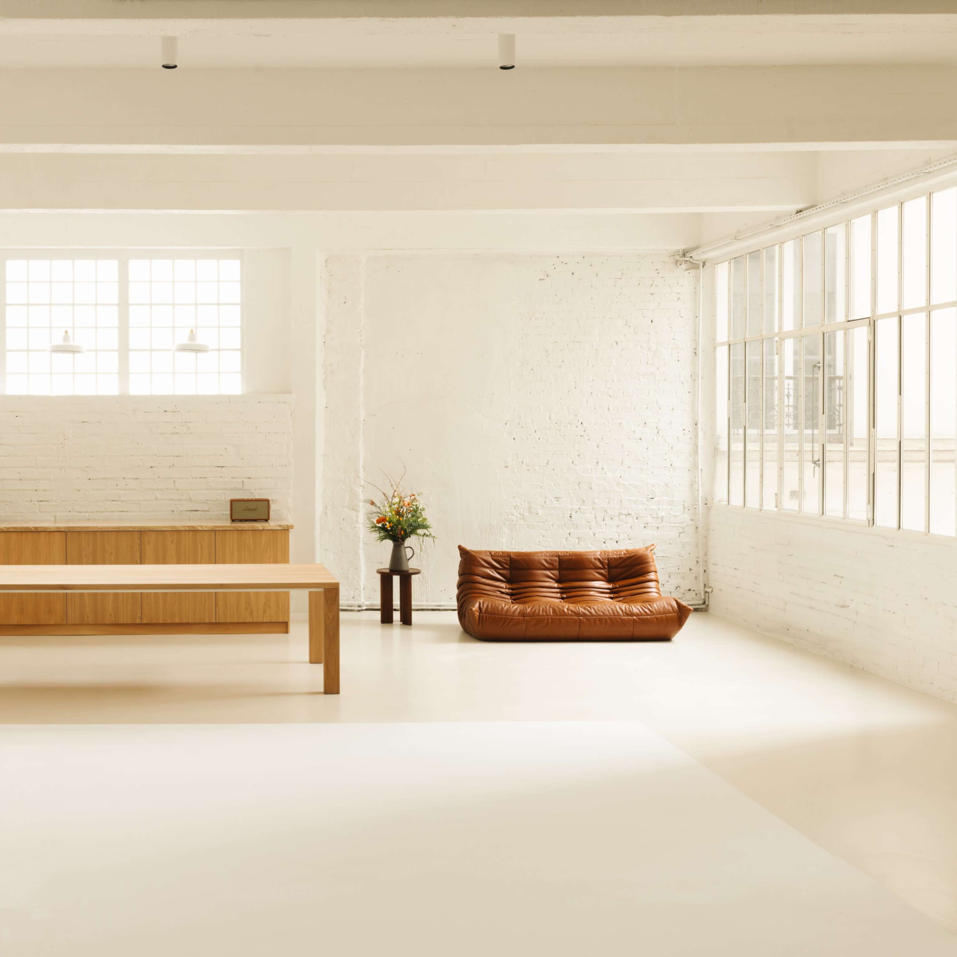 A spacious, minimalist interior with a wooden kitchen, a large dining table, and a brown sofa near a window with natural light.