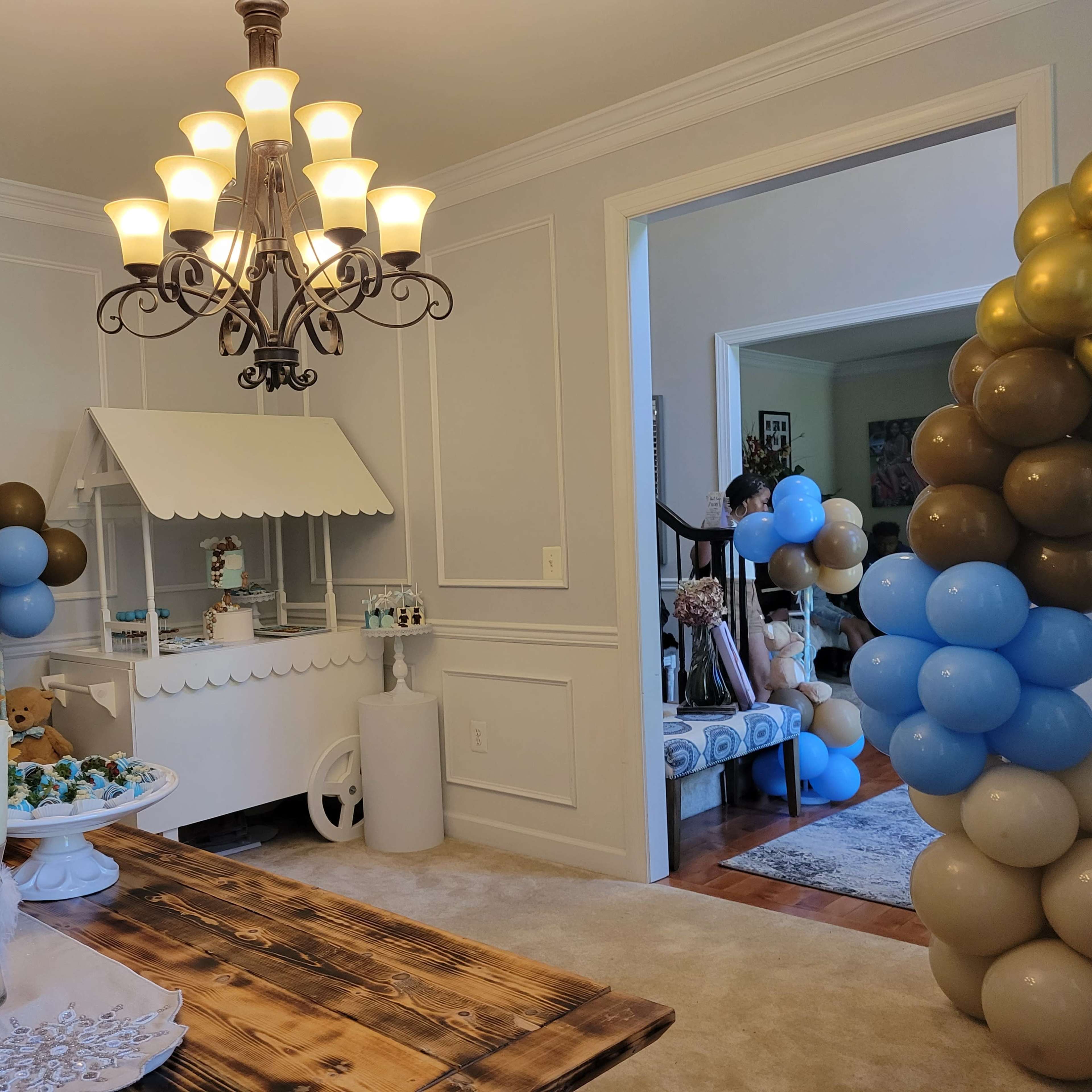 A decorated dining area features a dessert table with treats and a popcorn machine, surrounded by colorful balloons and a chandelier overhead.