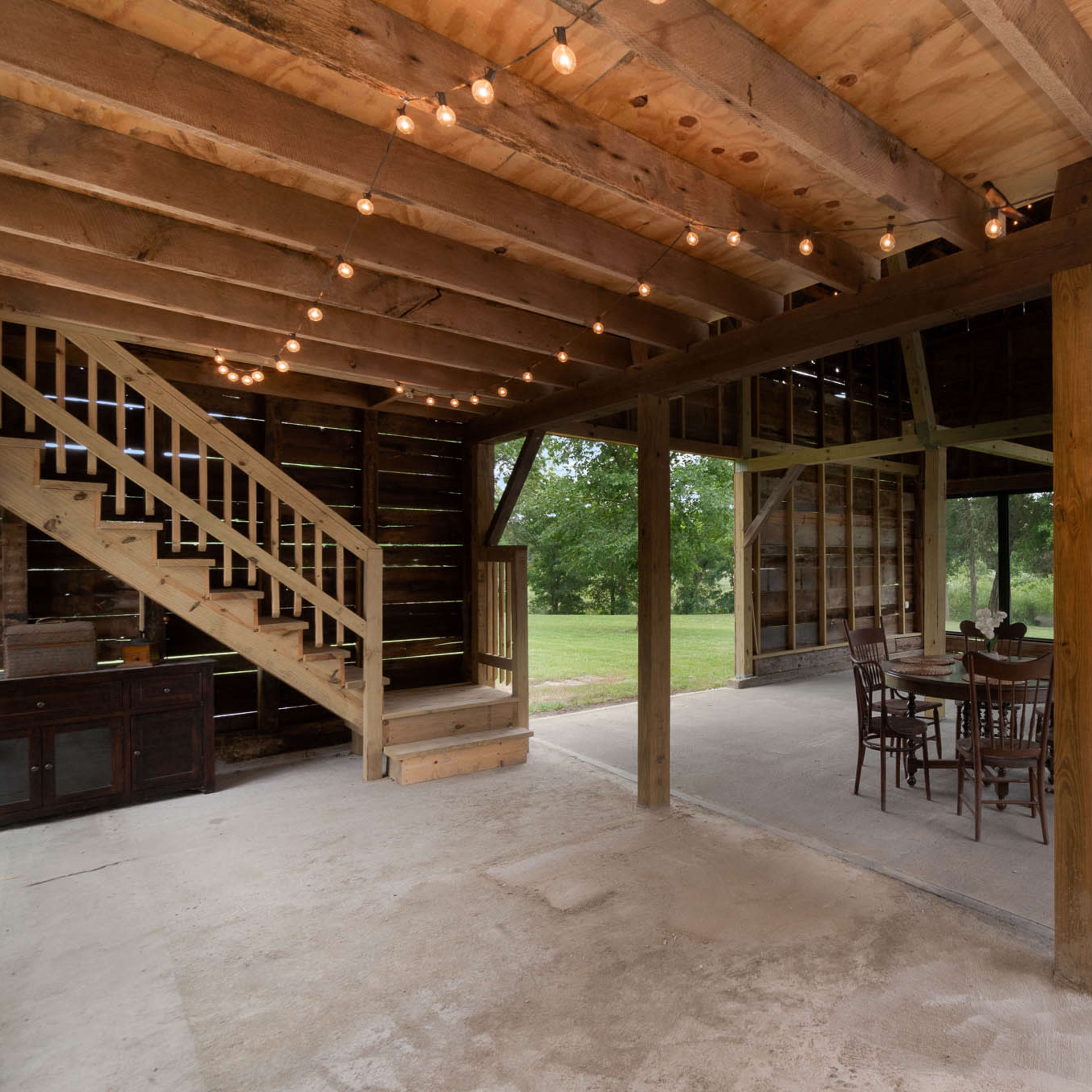 The interior of a rustic, open space features wooden beams, a staircase, a dining table with chairs, and large windows that overlook a green landscape.