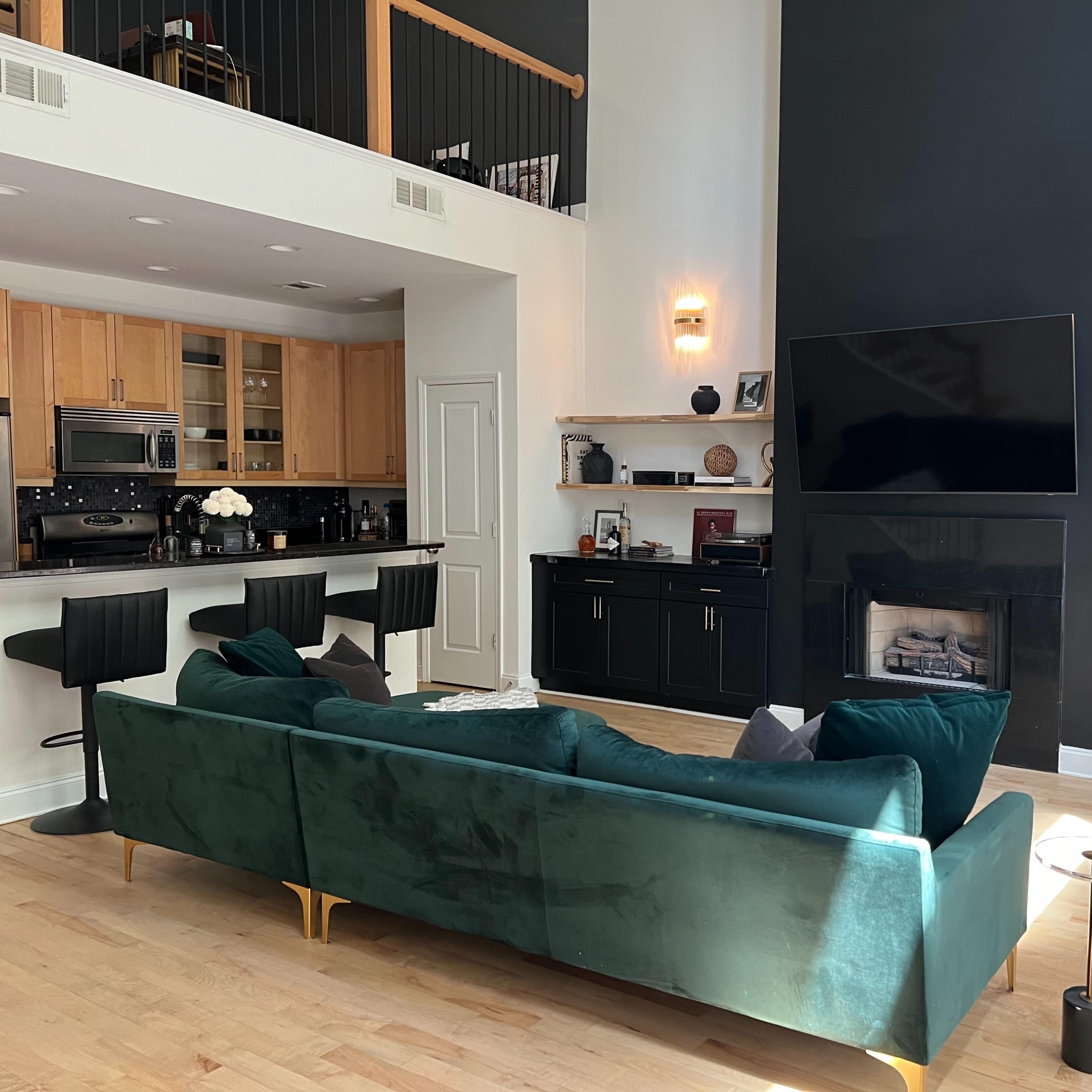 A modern living room with a green sectional sofa, a kitchen area with bar stools, and a minimalist black fireplace beneath a large flat-screen TV.