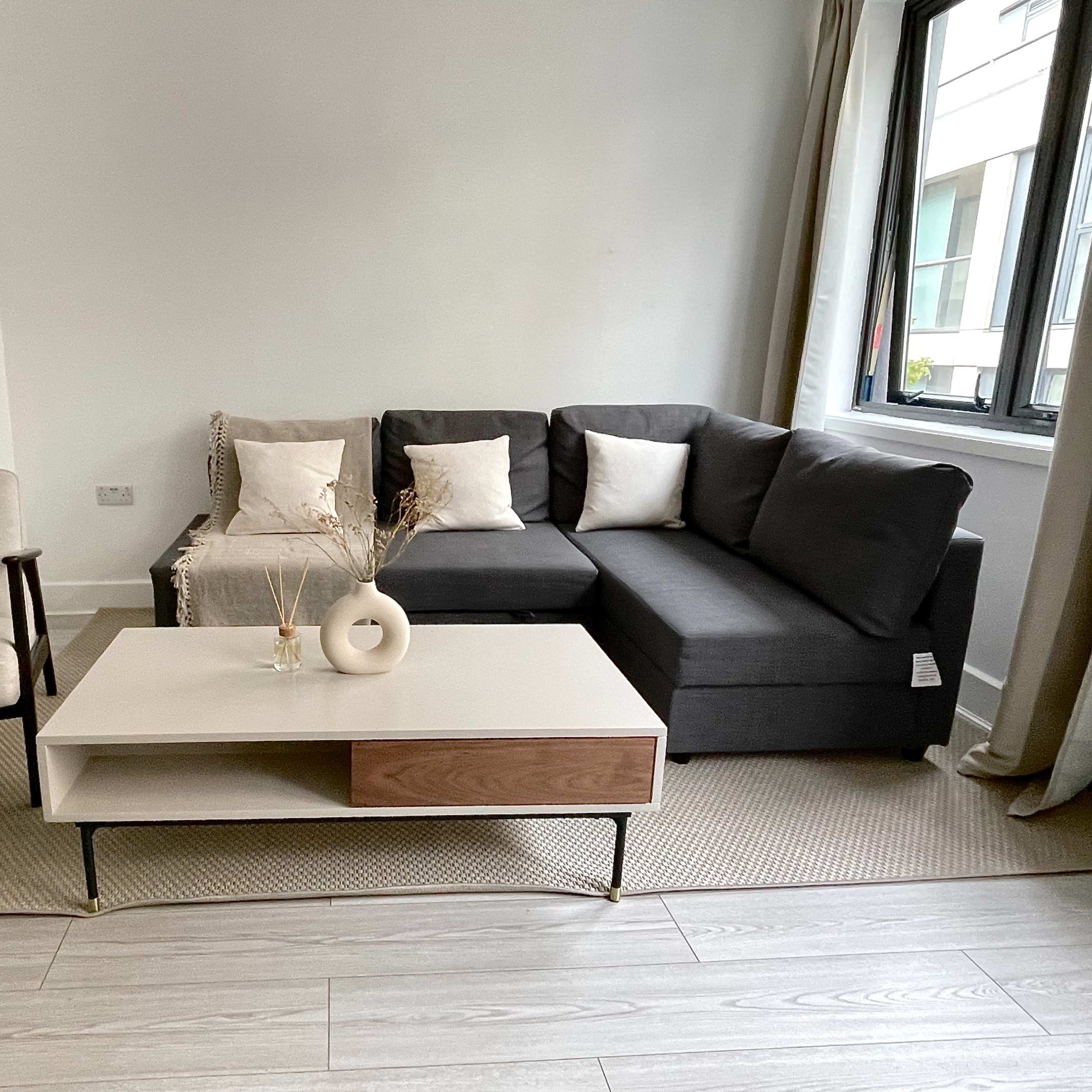 A modern living room features a dark sectional sofa, a light coffee table with a wooden drawer, and an armchair beside it.
