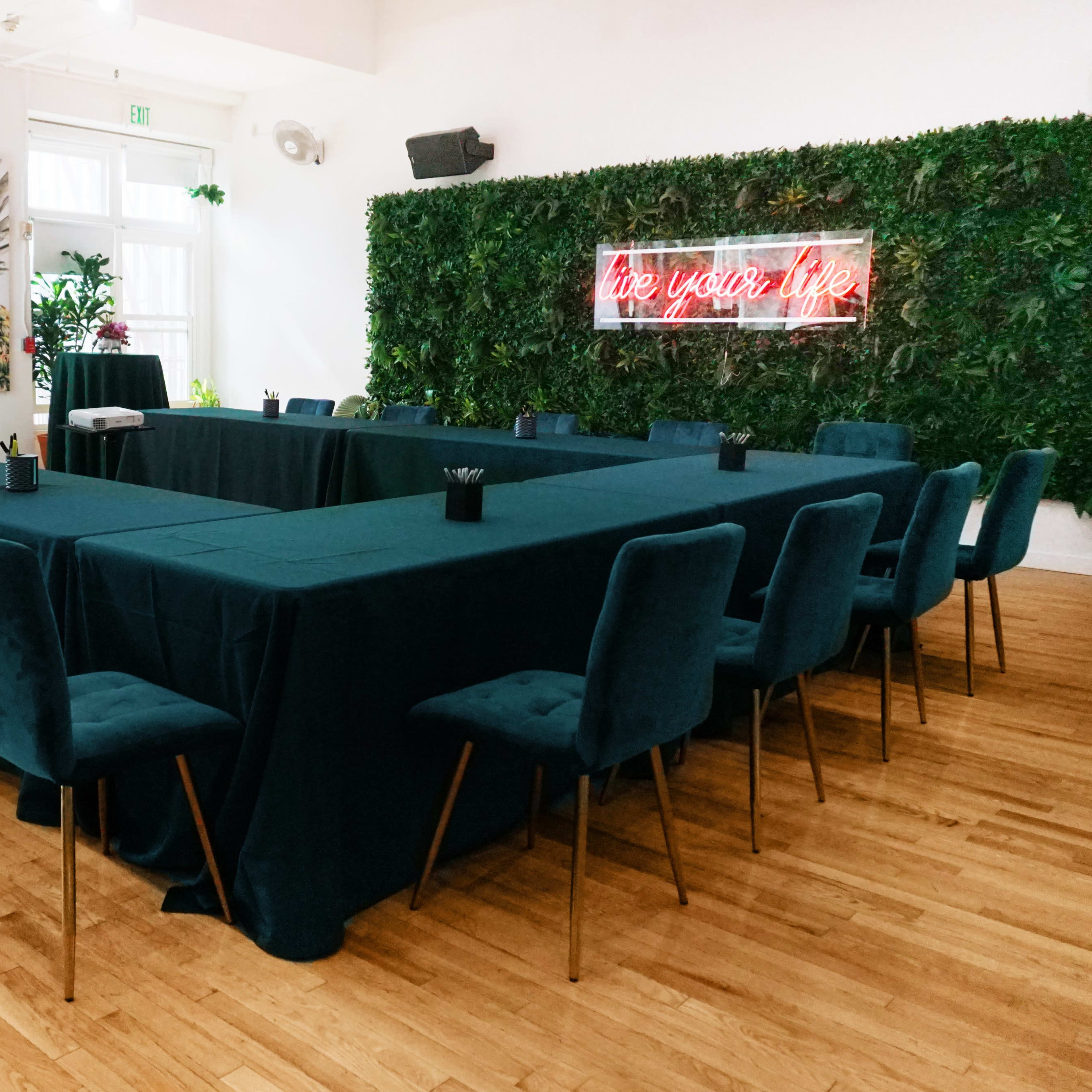 A rectangular conference room setup features a large table with teal linens surrounded by chairs, with a neon sign reading "live your life" and a wall of greenery in the background.