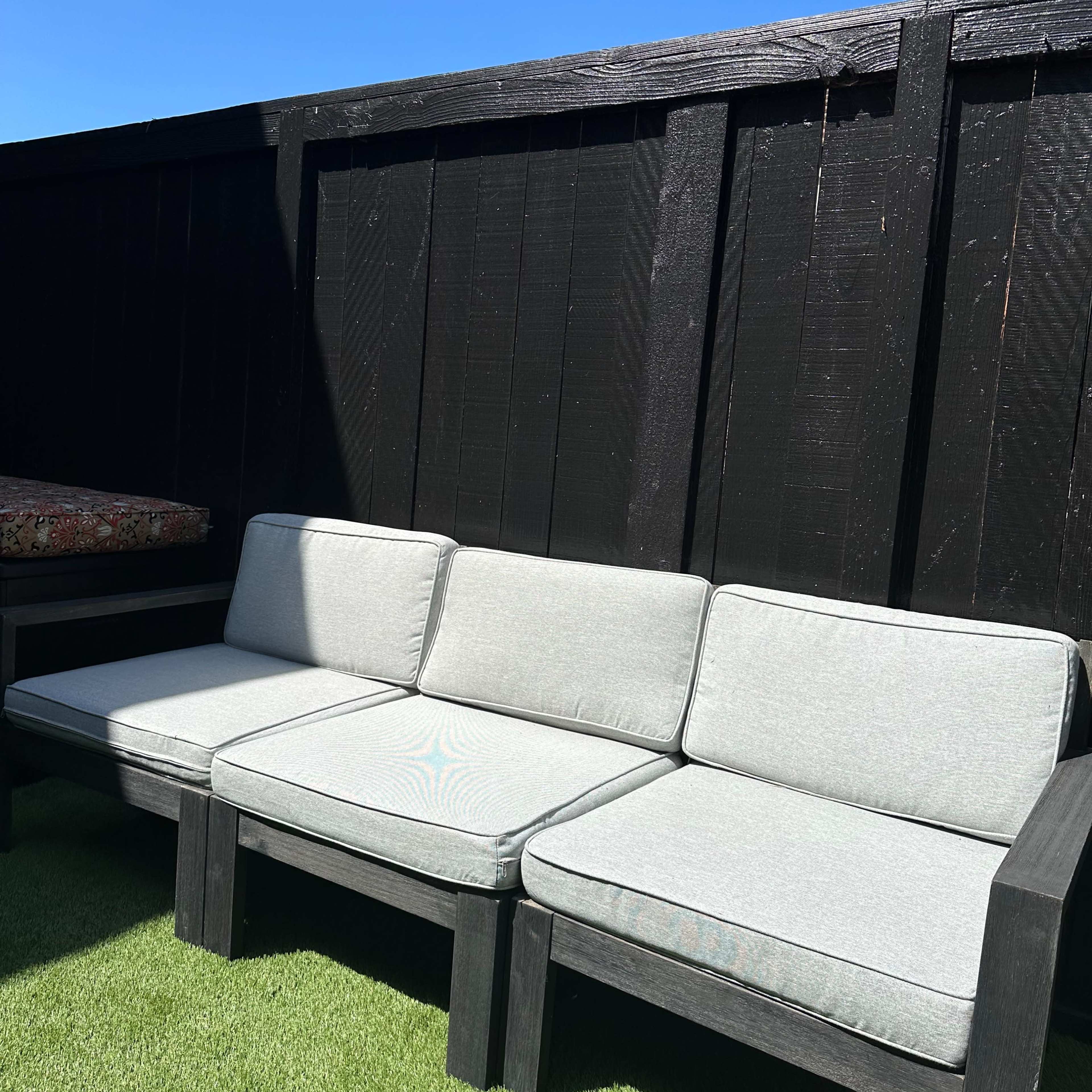A three-seat outdoor couch with gray cushions is positioned against a black wooden fence on a green lawn.
