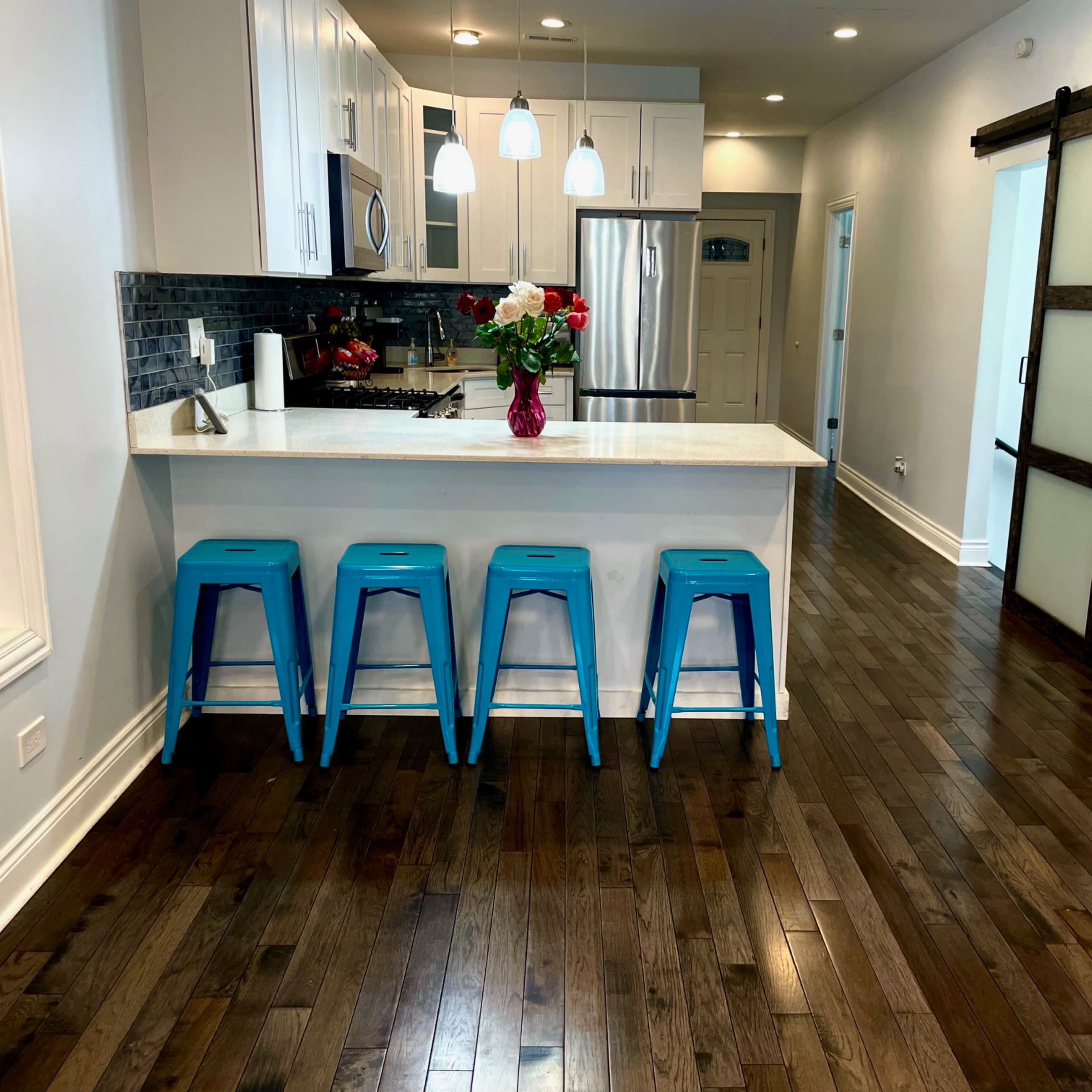 A modern kitchen features a white countertop with blue bar stools, stainless steel appliances, and hardwood flooring.