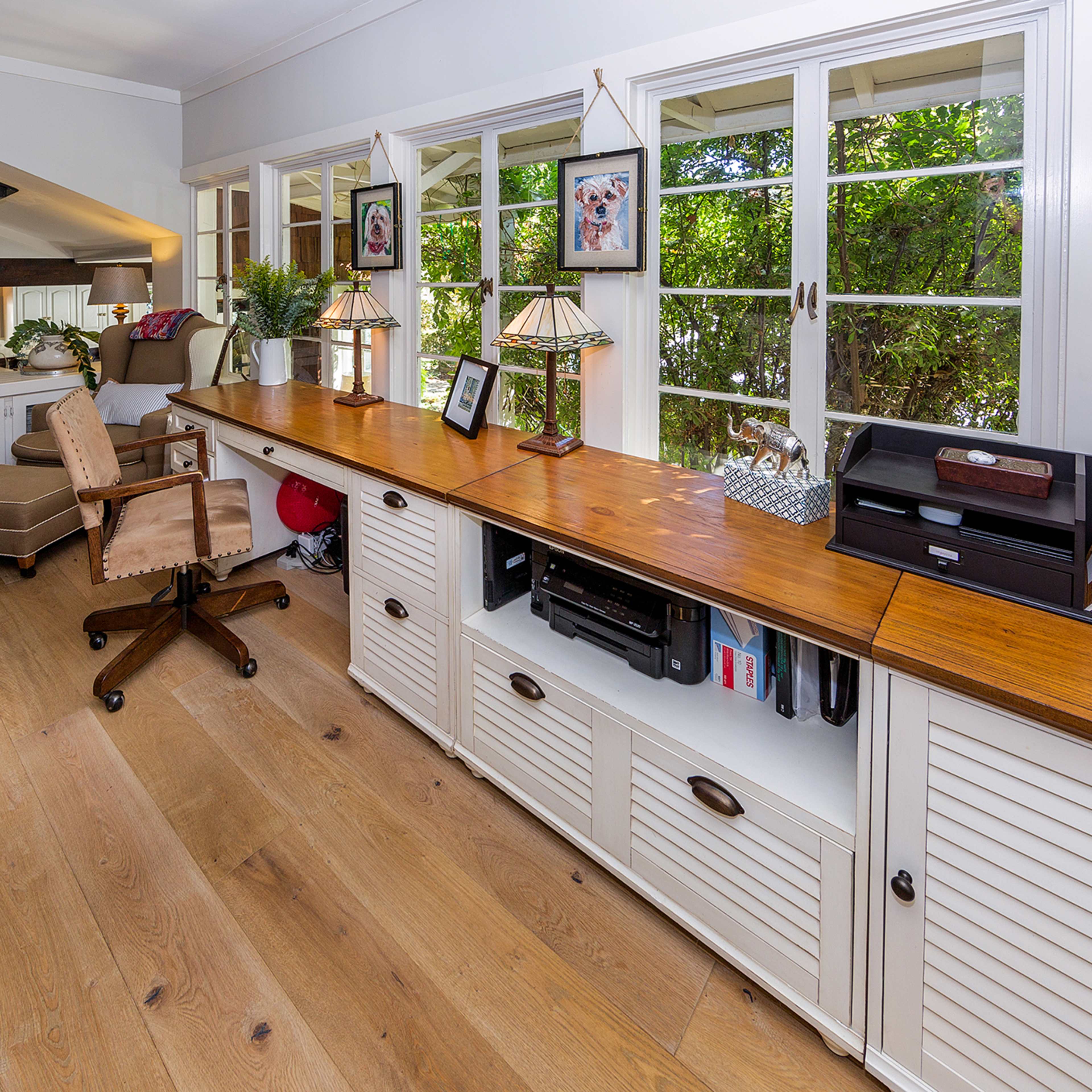 A well-lit room featuring a wooden sideboard, a plush chair, and large windows with greenery outside.