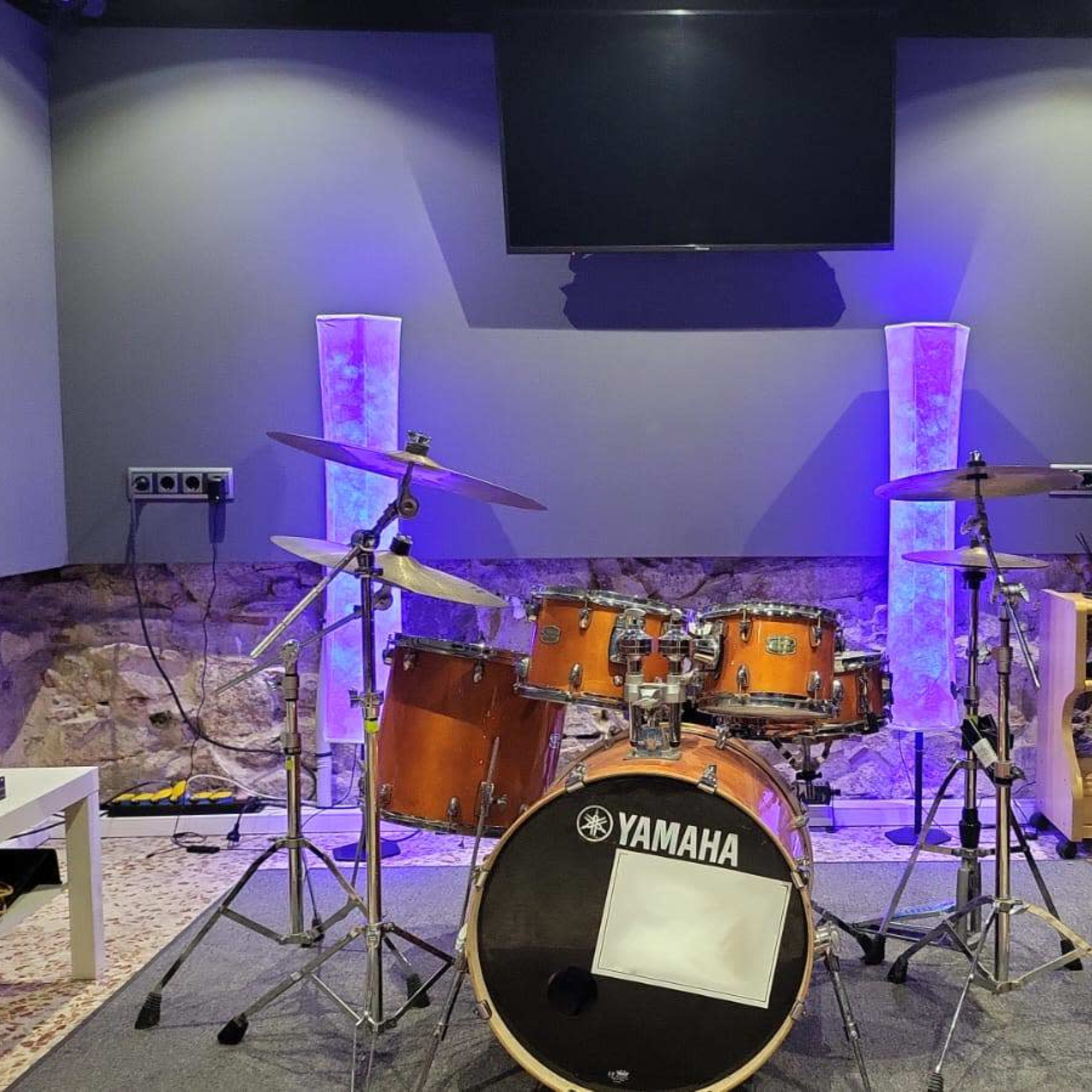A music studio features a drum set, an amplifier, and a television screen, with purple ambient lighting and a stone wall backdrop.