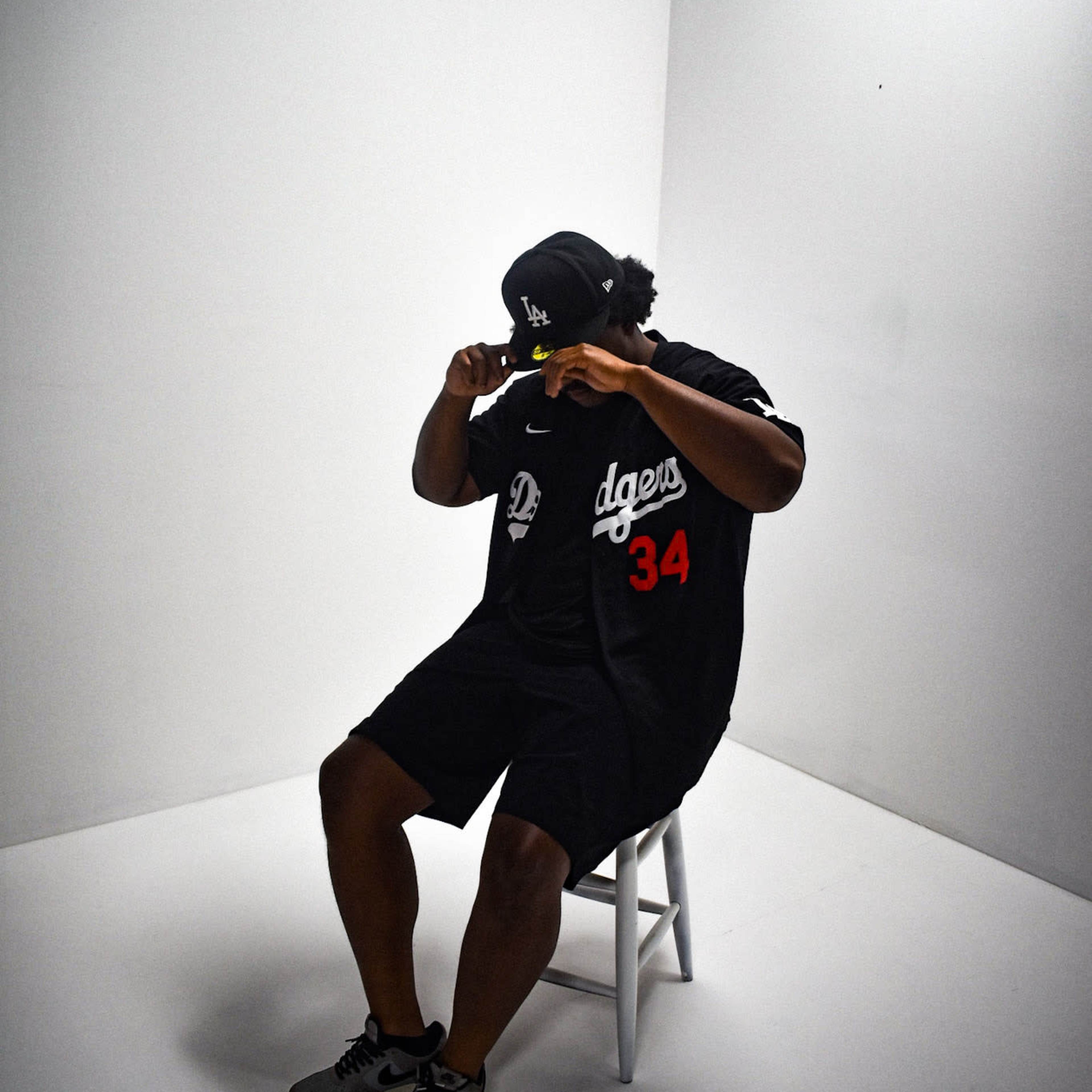A person wearing a Dodgers jersey and cap sits on a stool in a plain, well-lit studio.