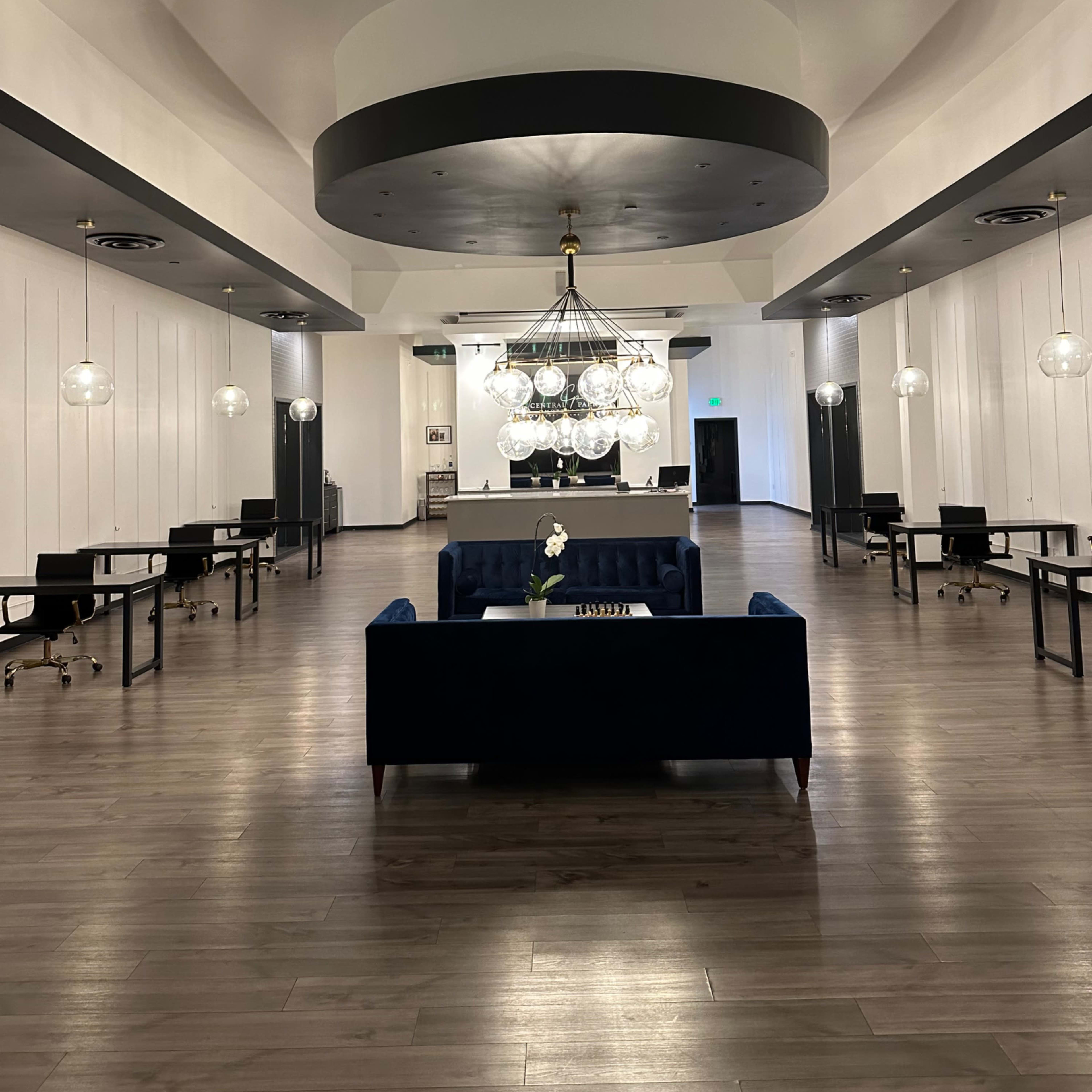 A spacious lobby area featuring a central chandelier, a blue sofa, and several desks arranged along the walls.