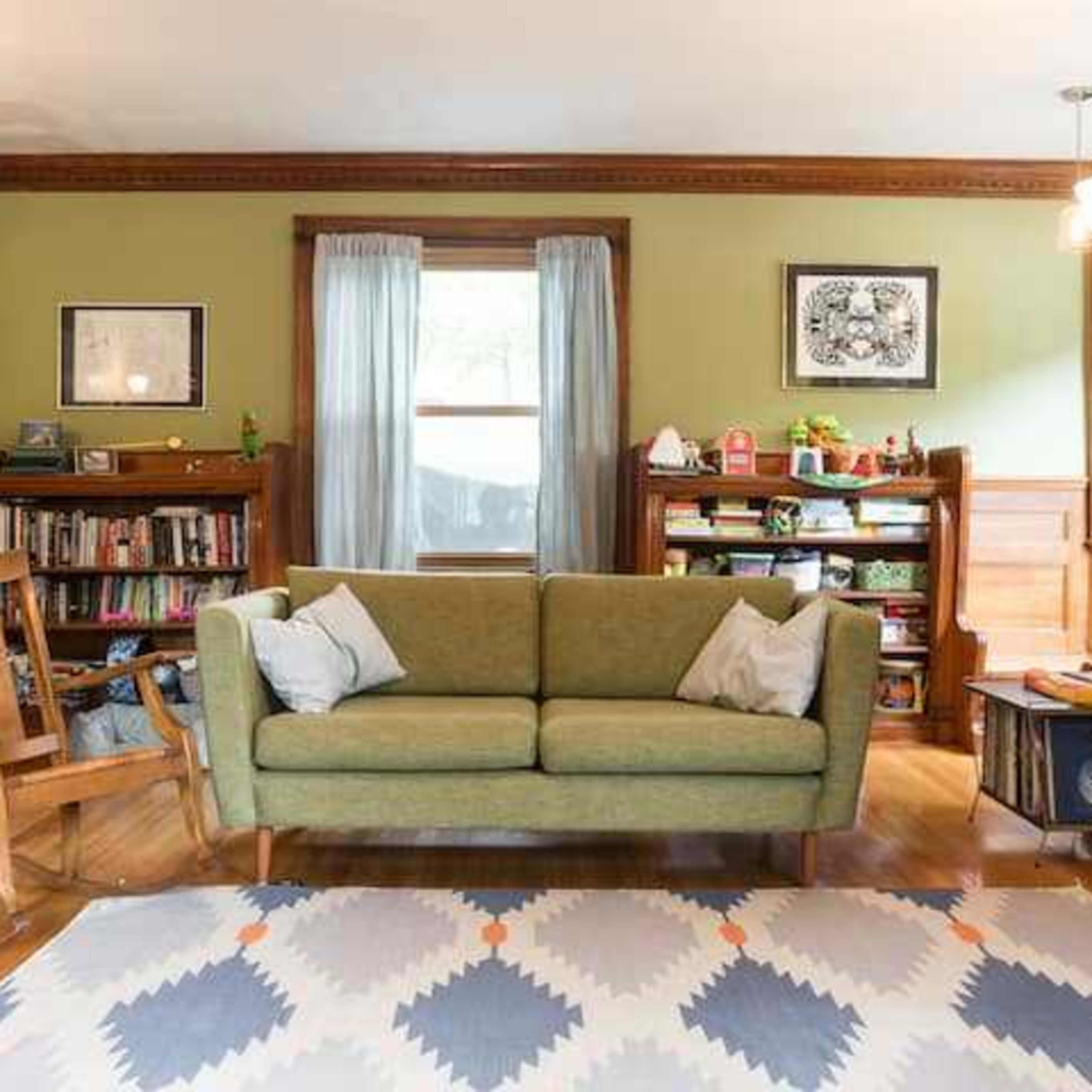 The living room features a green sofa, a wooden rocking chair, a bookshelf with various items, and a fireplace with a mantel, all set against a backdrop of light green walls and wooden trim.
