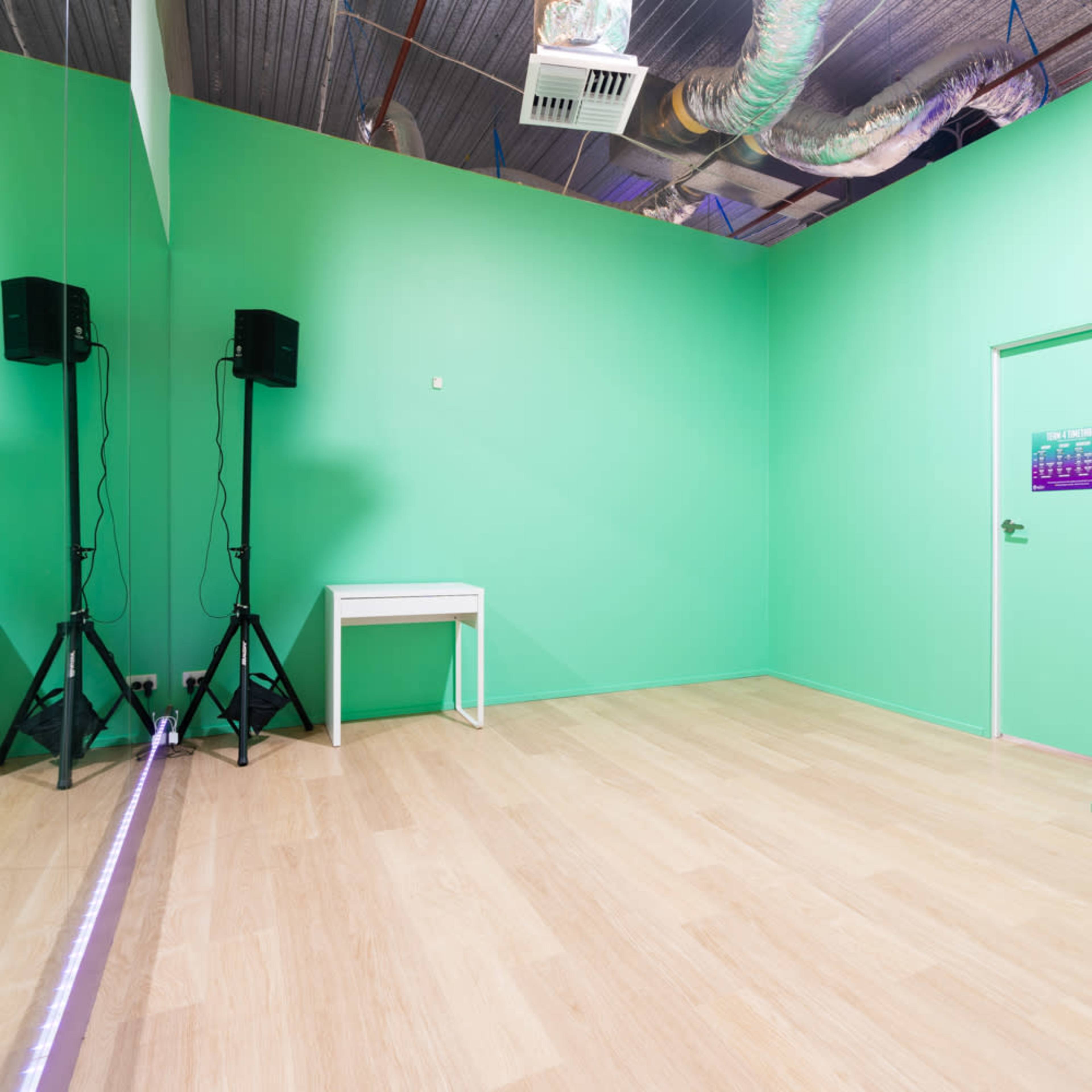 A room with green walls, wooden flooring, a mirrored wall, two speakers on stands, and a door.