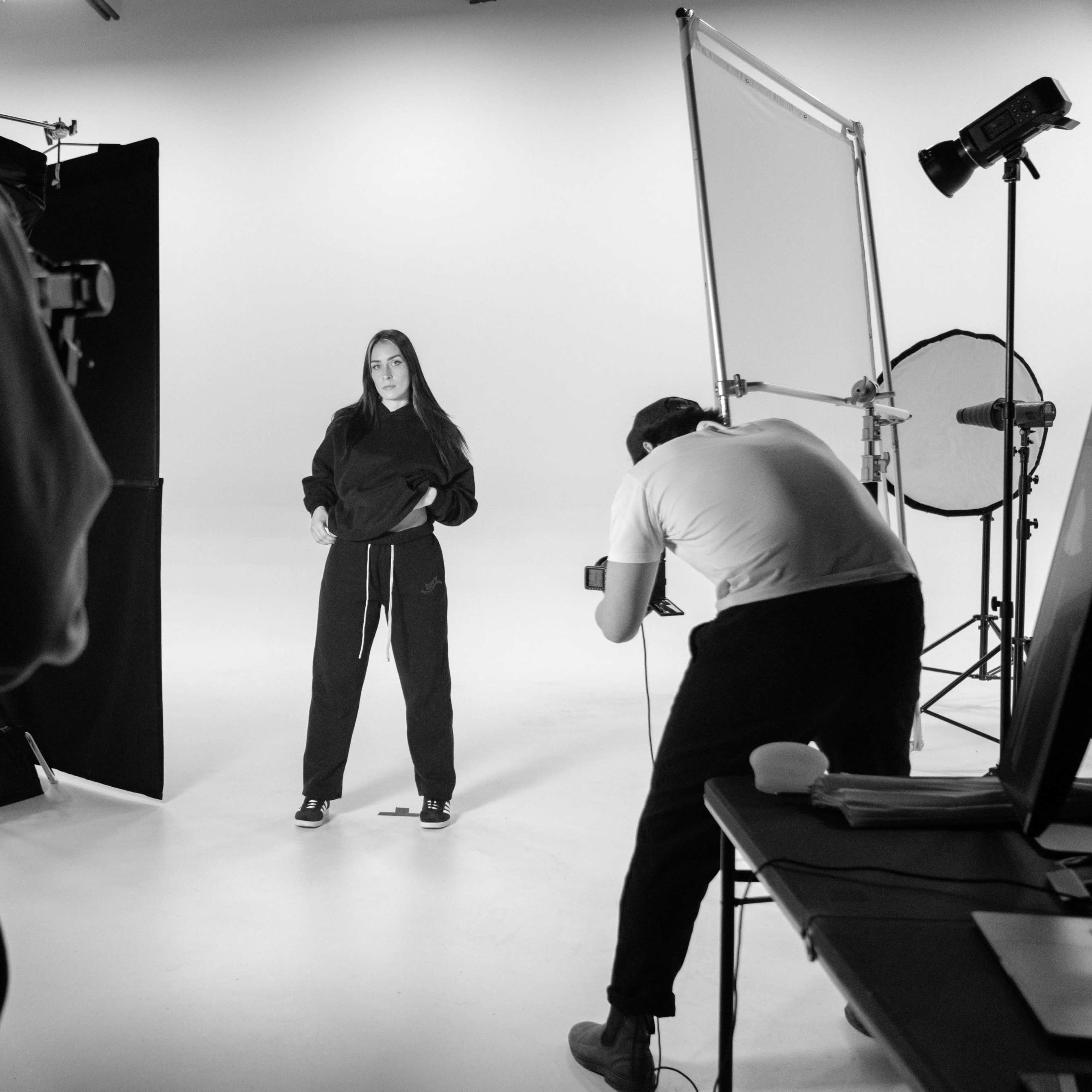A person in a hooded outfit poses for a photo while another individual sets up a camera in a studio.