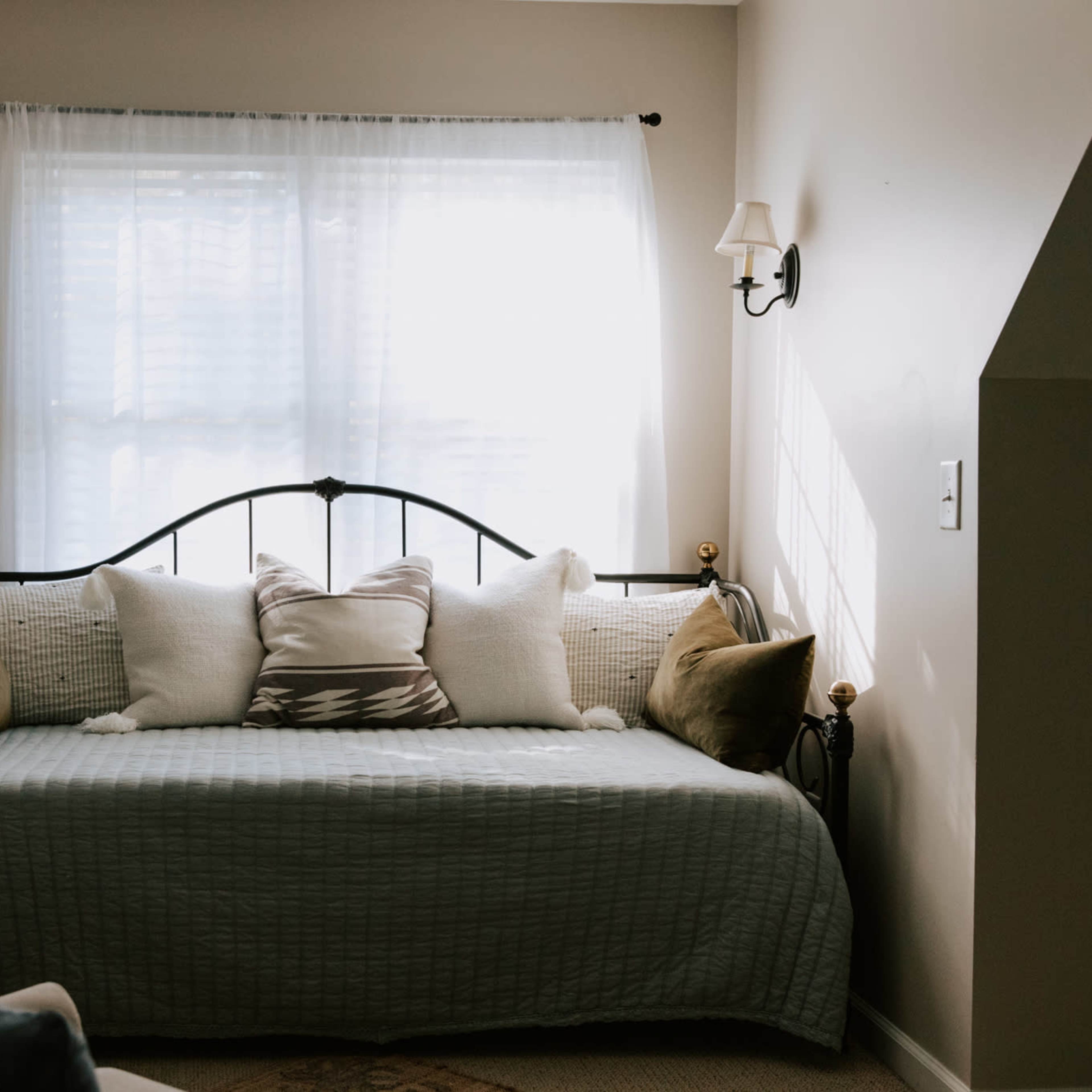 A daybed with decorative pillows is positioned against a wall under a window with sheer curtains in a softly lit room.