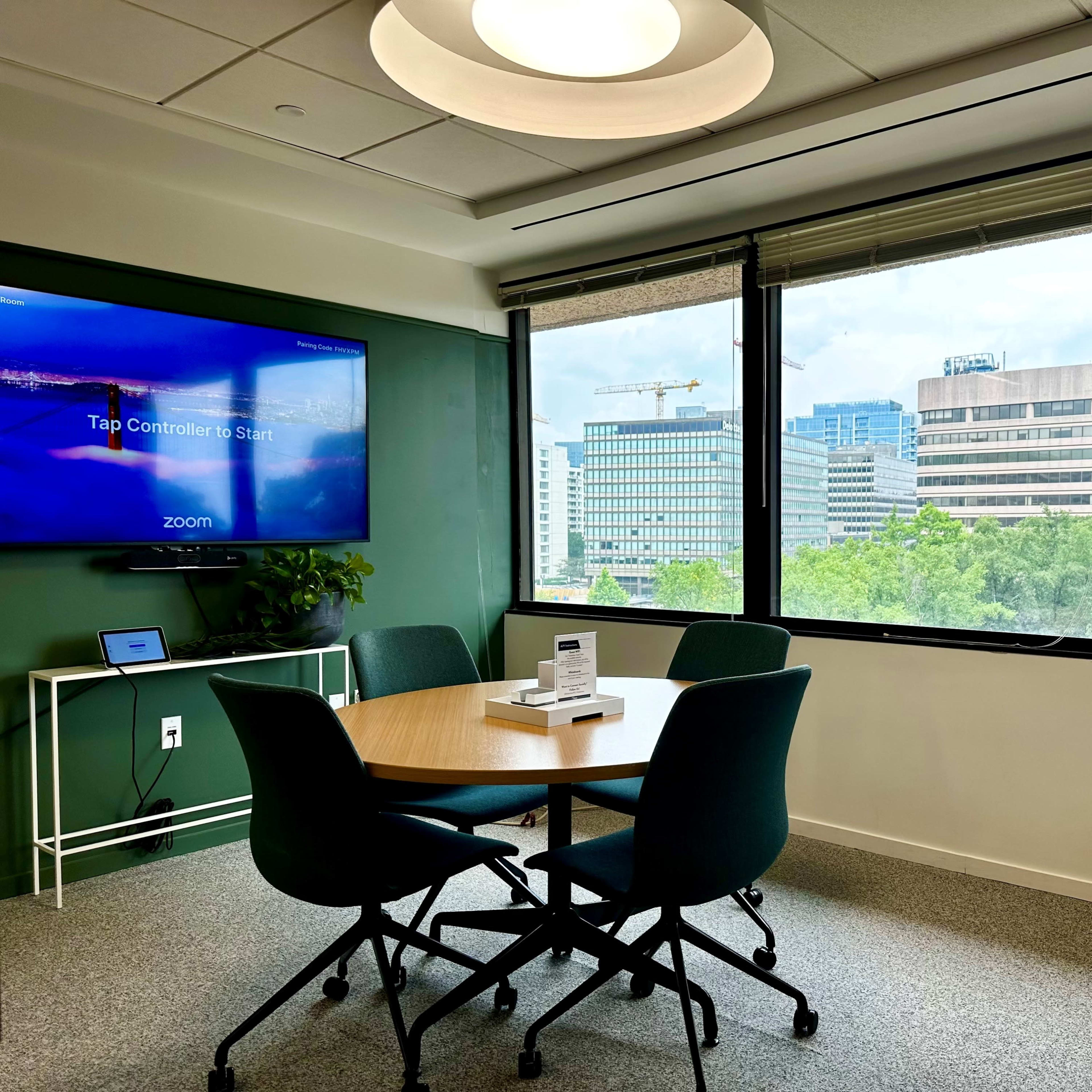 A modern meeting room with a circular table, four chairs, a screen displaying "Tap Controller to Start," and large windows that overlook a cityscape.