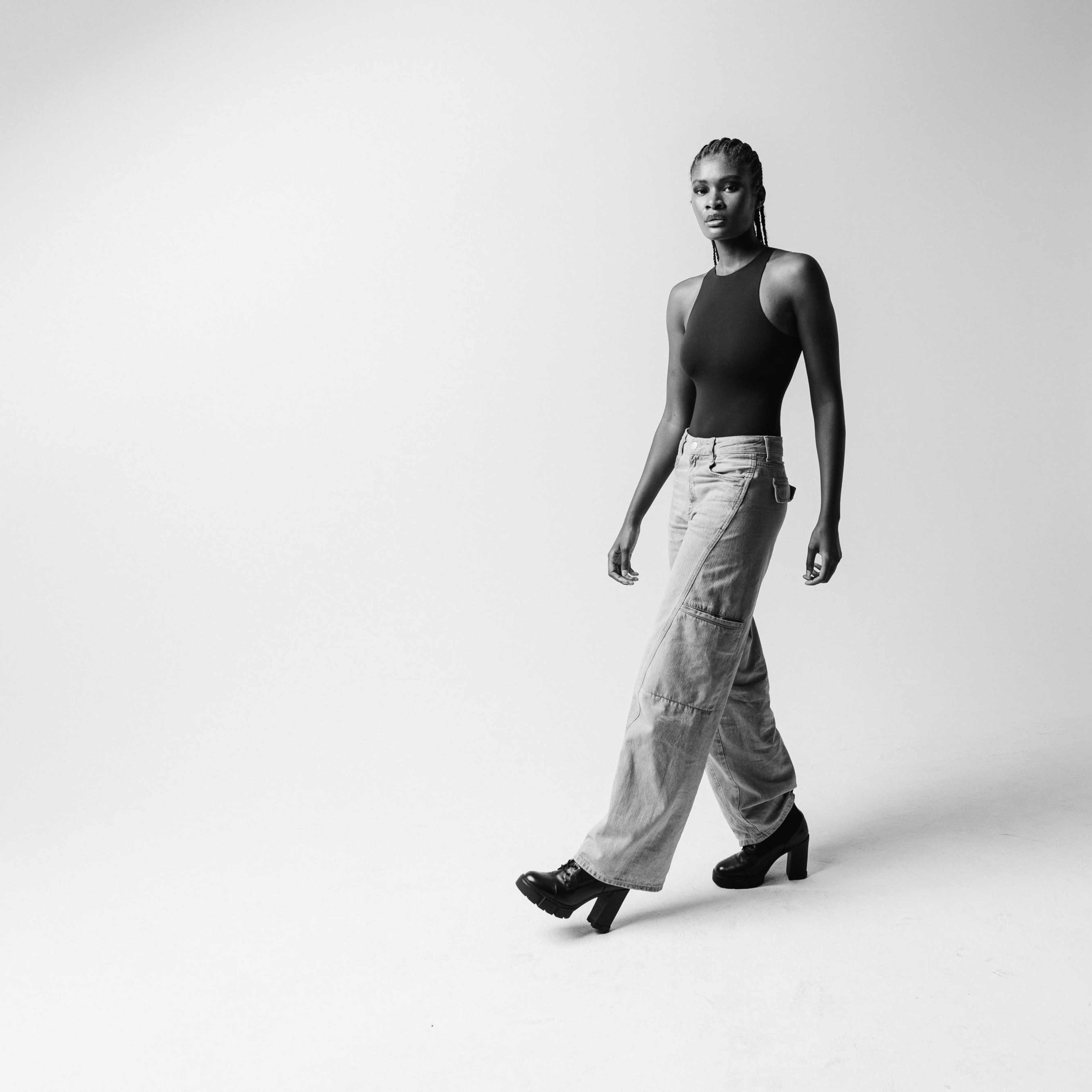 A person walks confidently in a minimalist studio space, wearing a sleeveless top and wide-legged pants.