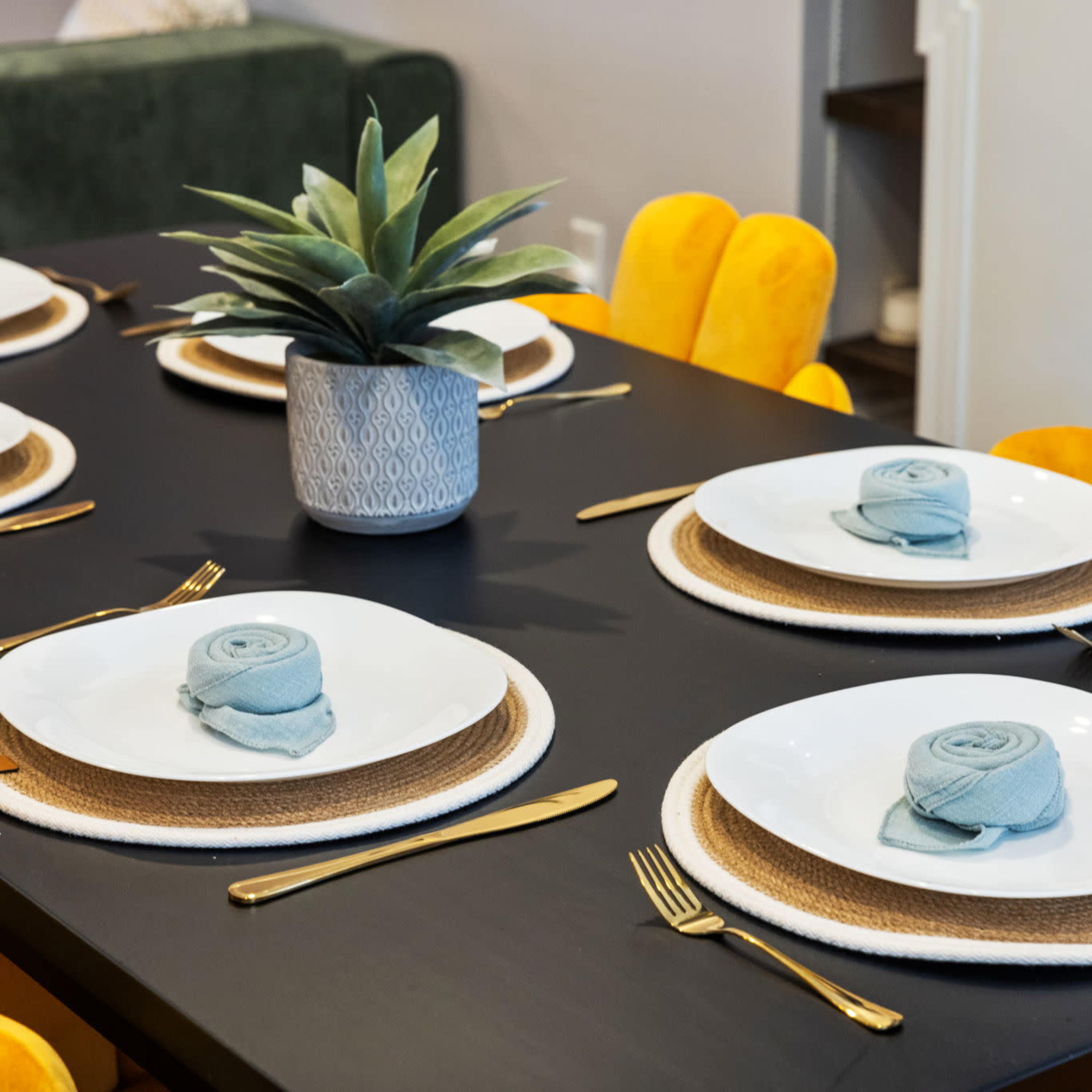 A dining table is set with six white plates on woven placemats, adorned with rolled blue napkins and a succulent centerpiece, surrounded by yellow chairs.