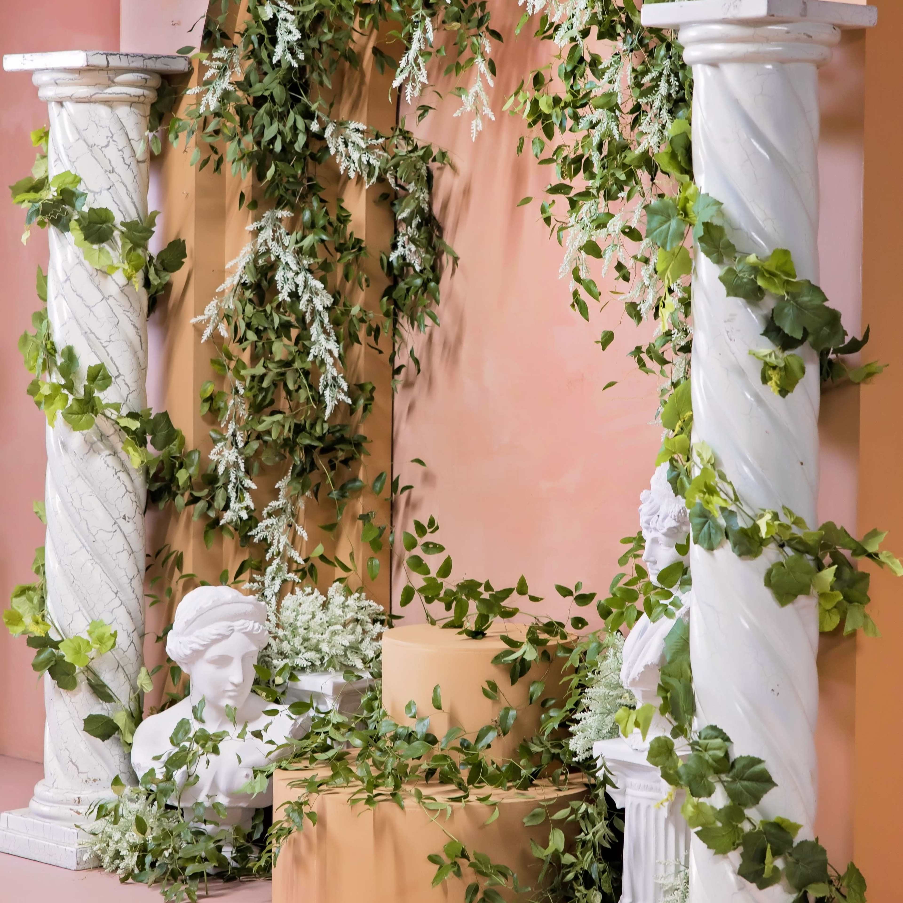 The scene features two white, fluted columns supporting a decorative arch adorned with greenery and floral arrangements, flanking a central pedestal surrounded by additional foliage and two white busts.