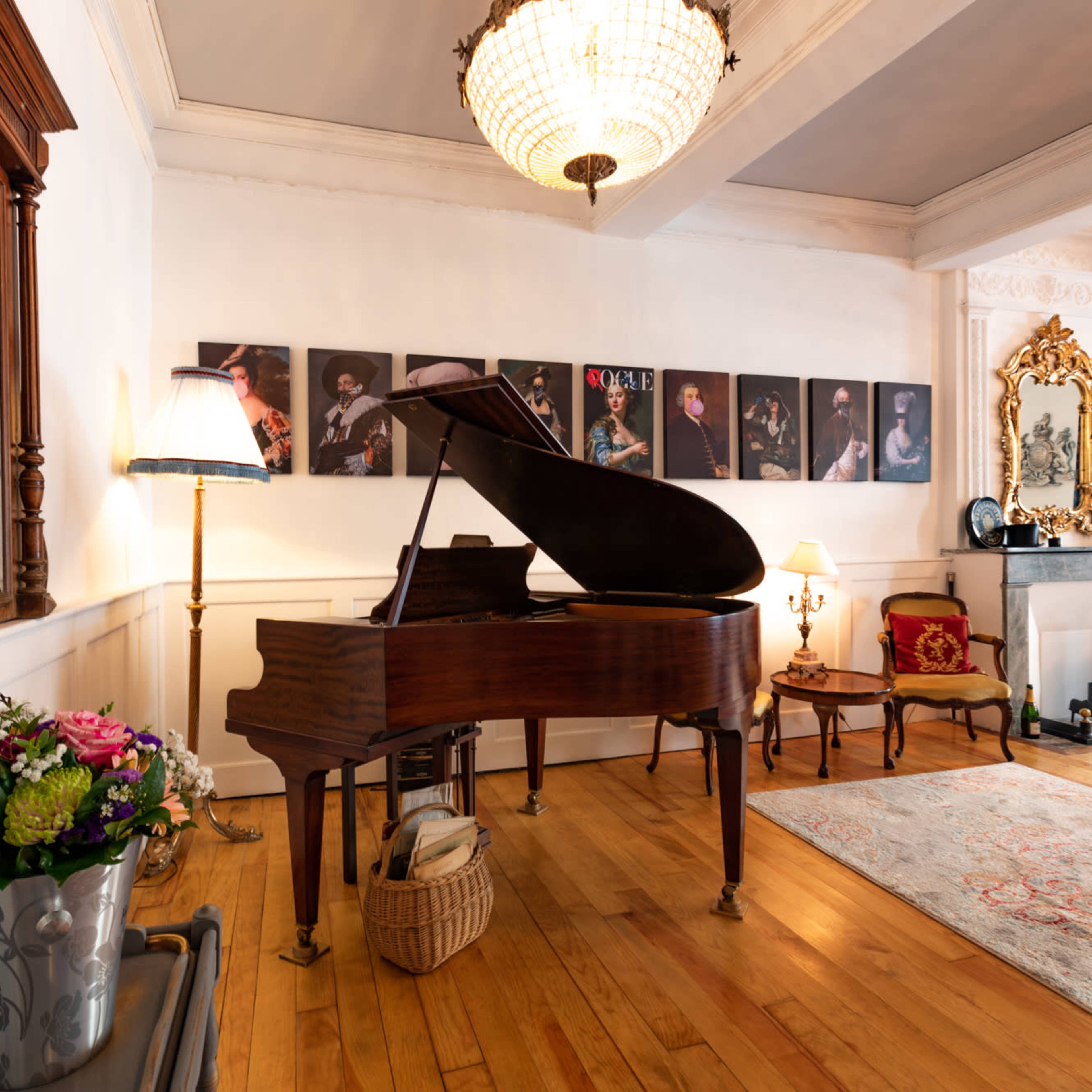 A grand piano is positioned in a room adorned with framed photographs, a large chandelier hangs overhead, and a rug covers the wooden floor.