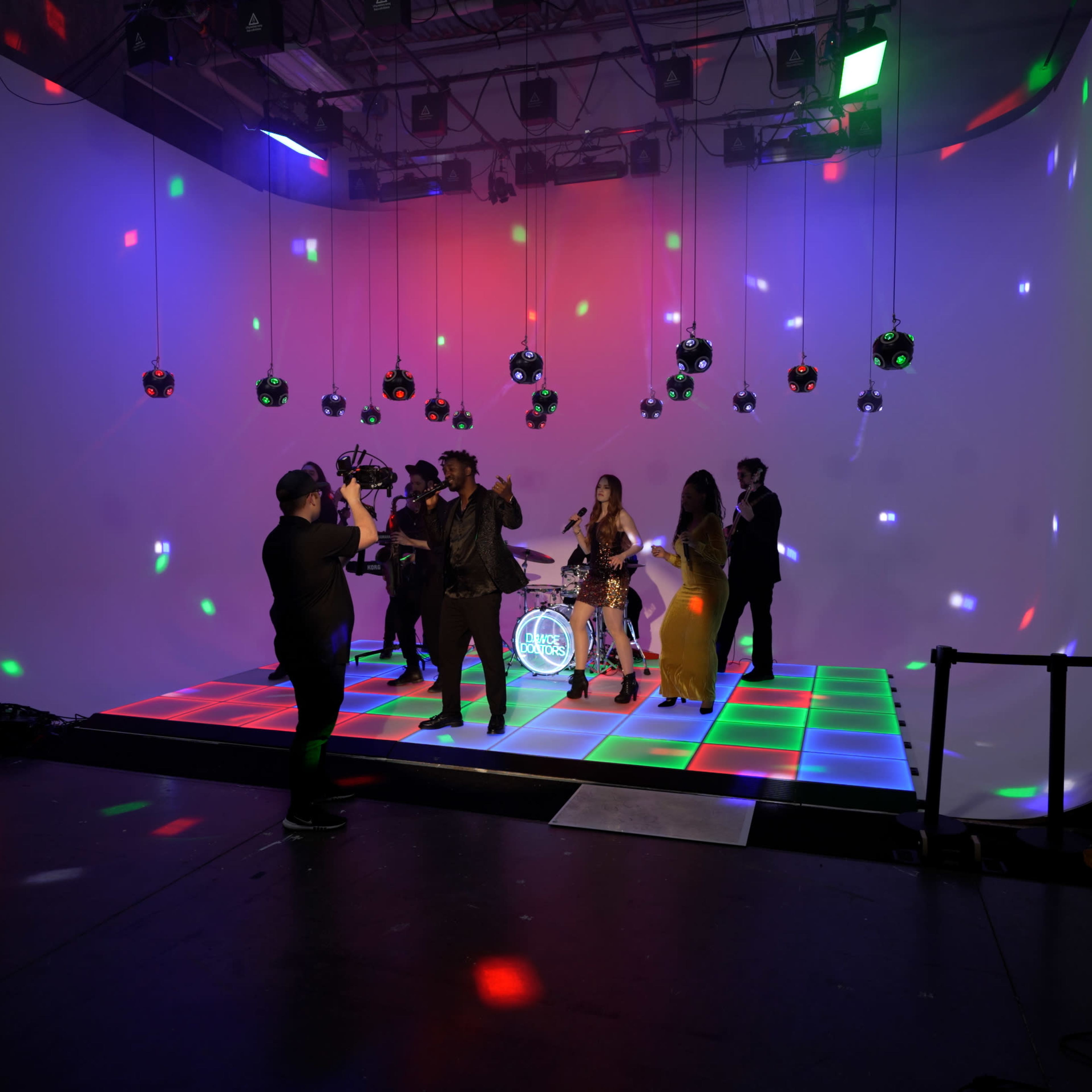 A group of musicians performs on a brightly lit stage with colorful lights and reflective decorations hanging overhead.