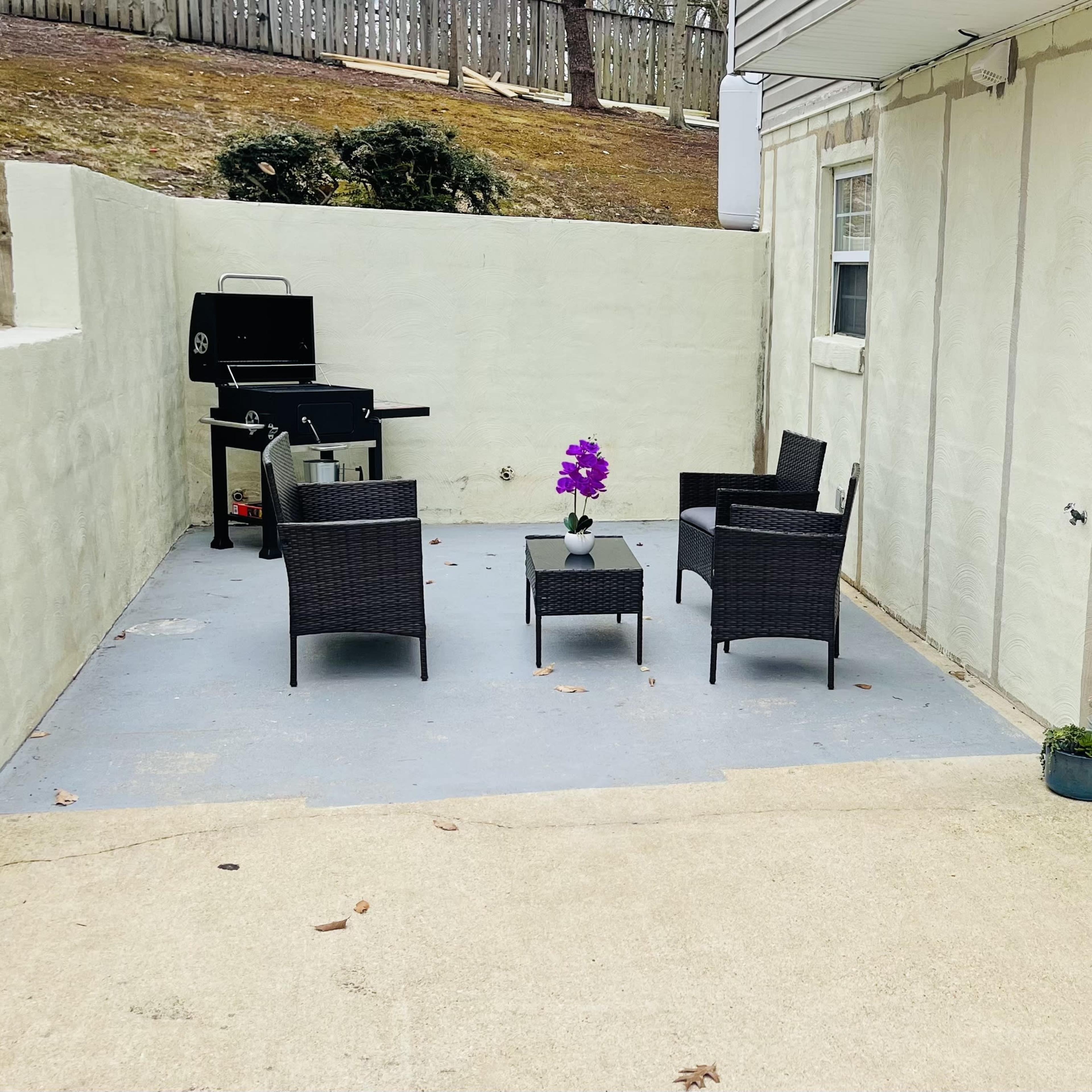 A small outdoor patio area features a set of four wicker chairs and a table, along with a grill against the wall.