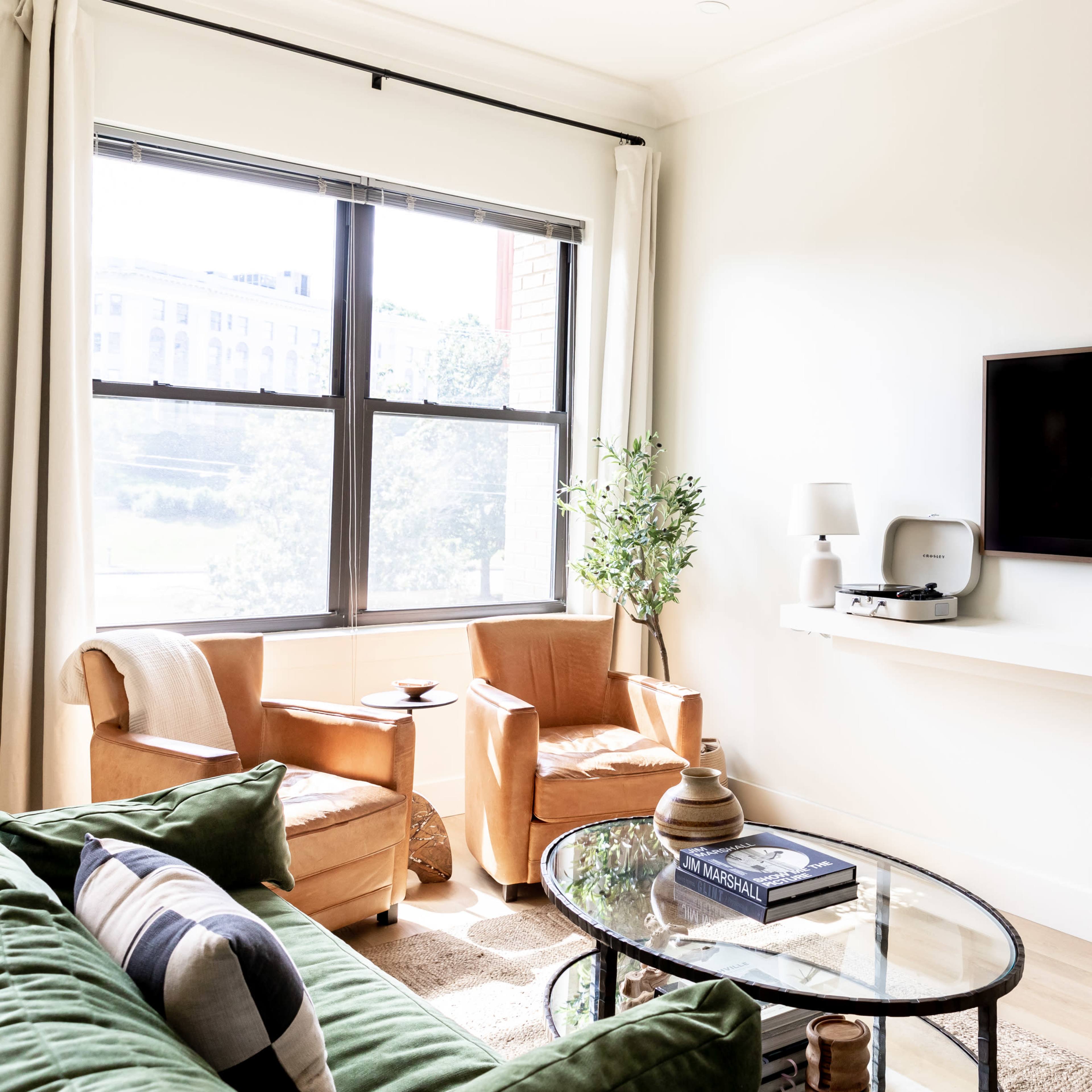A modern living room features a green sofa, two brown chairs, a coffee table, and a mounted television on a white wall.