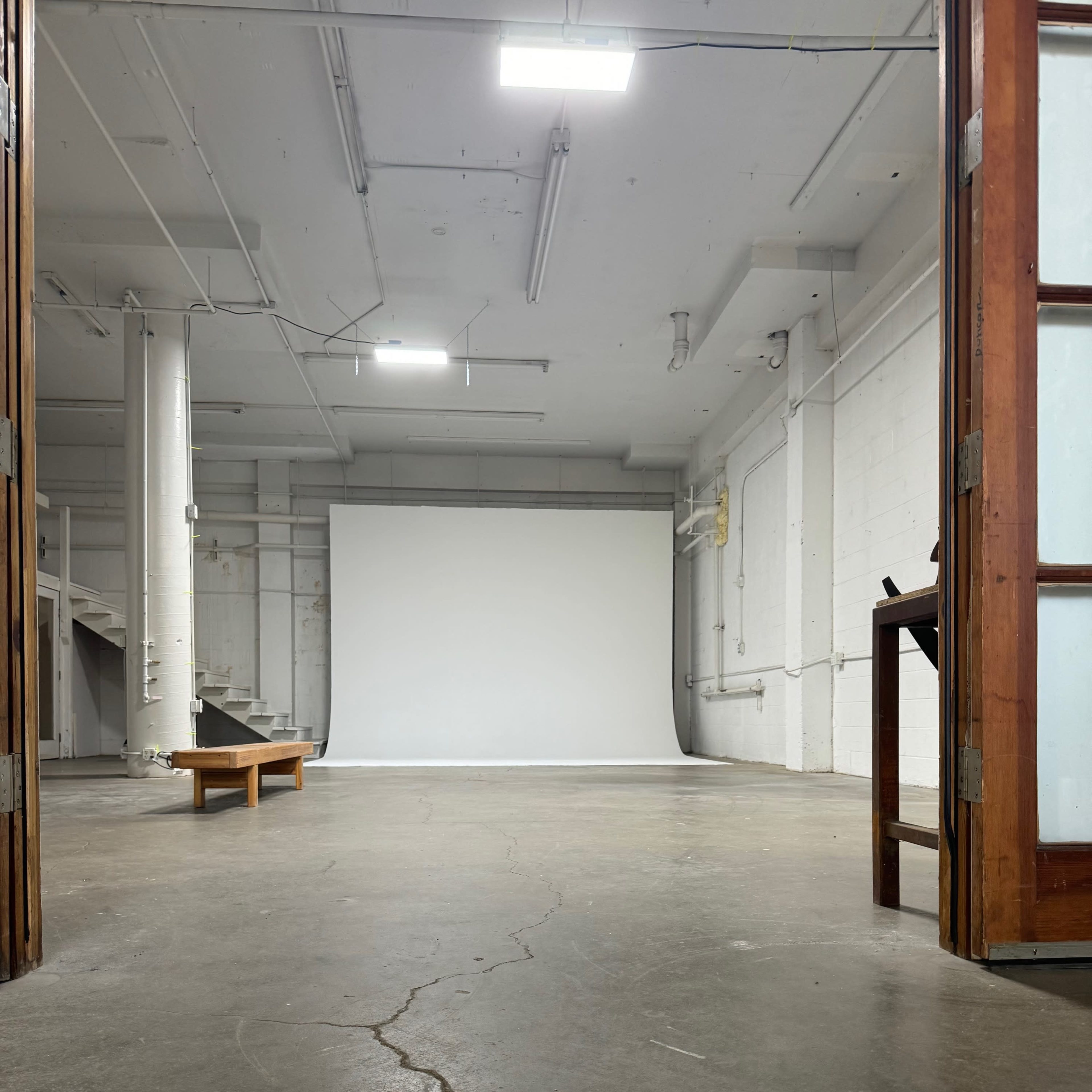 An empty studio space with a blank white backdrop at the far end and a wooden bench in the foreground.