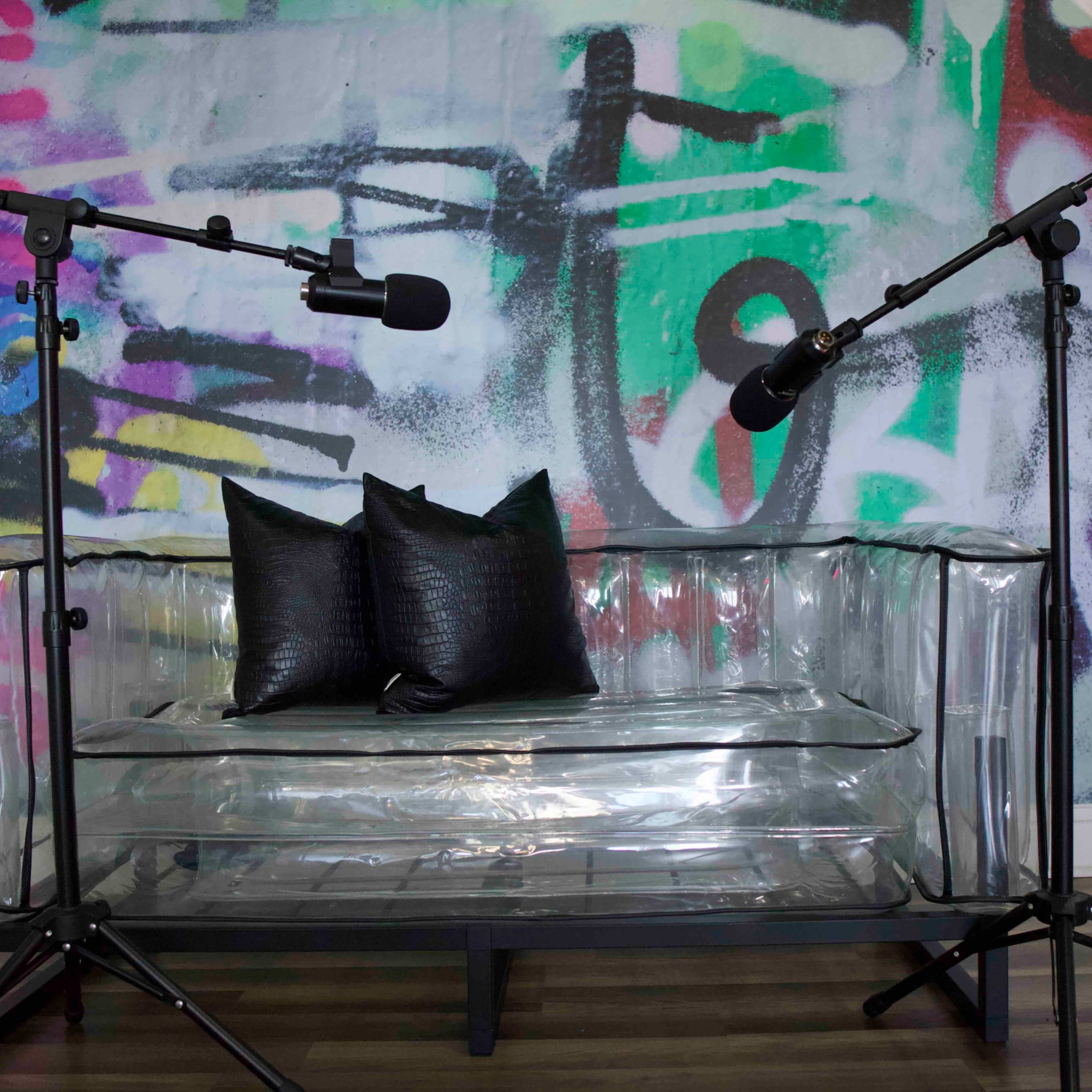 A transparent acrylic couch with two black pillows is positioned in front of a colorful graffiti-style wall, flanked by two microphone stands.