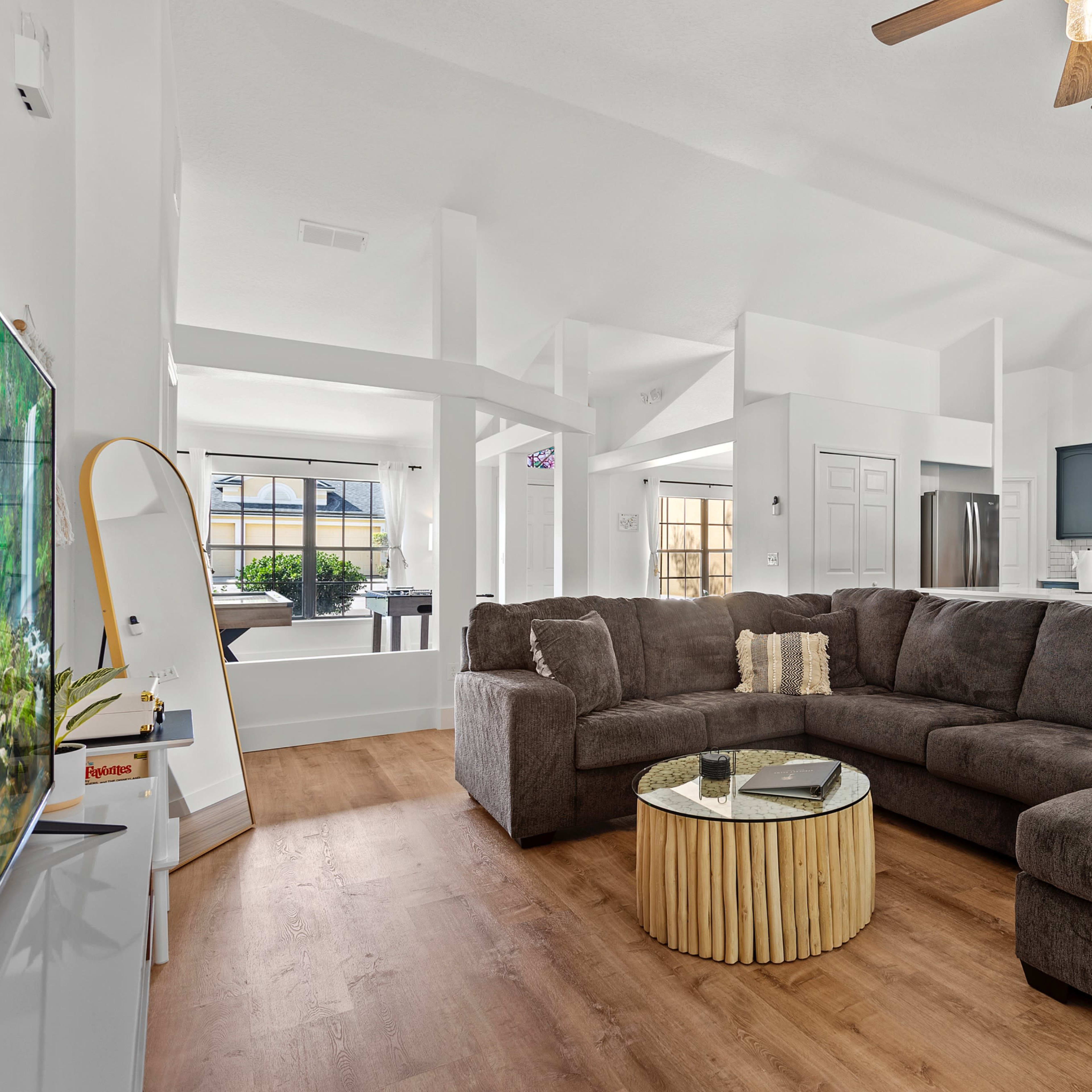 A spacious living room features a large, dark sectional sofa, a round coffee table made of wood, and a flat-screen TV on a white wall, with an open kitchen visible in the background.