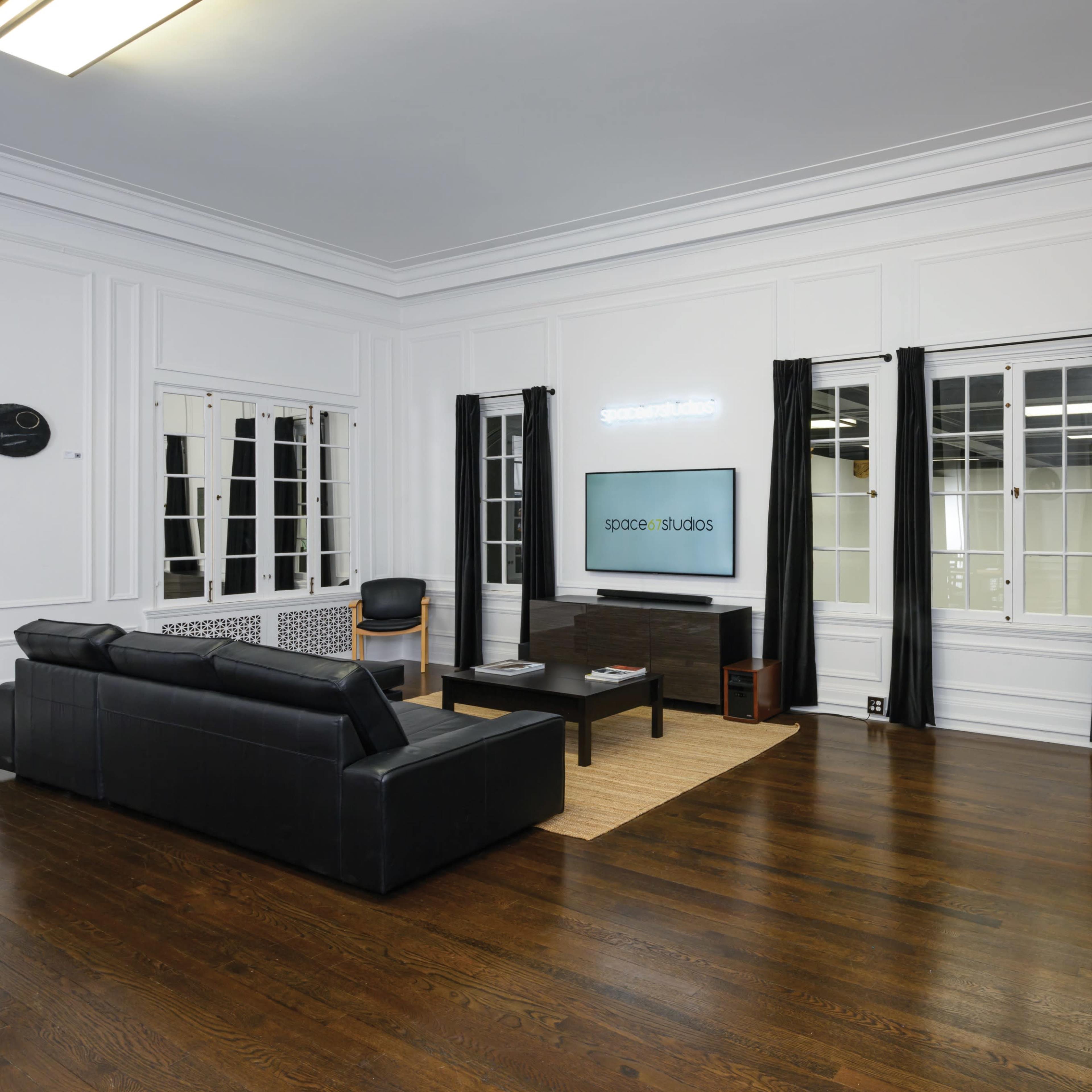 The room features dark hardwood flooring, white walls, large windows with black curtains, and modern furniture including a black couch and chair.
