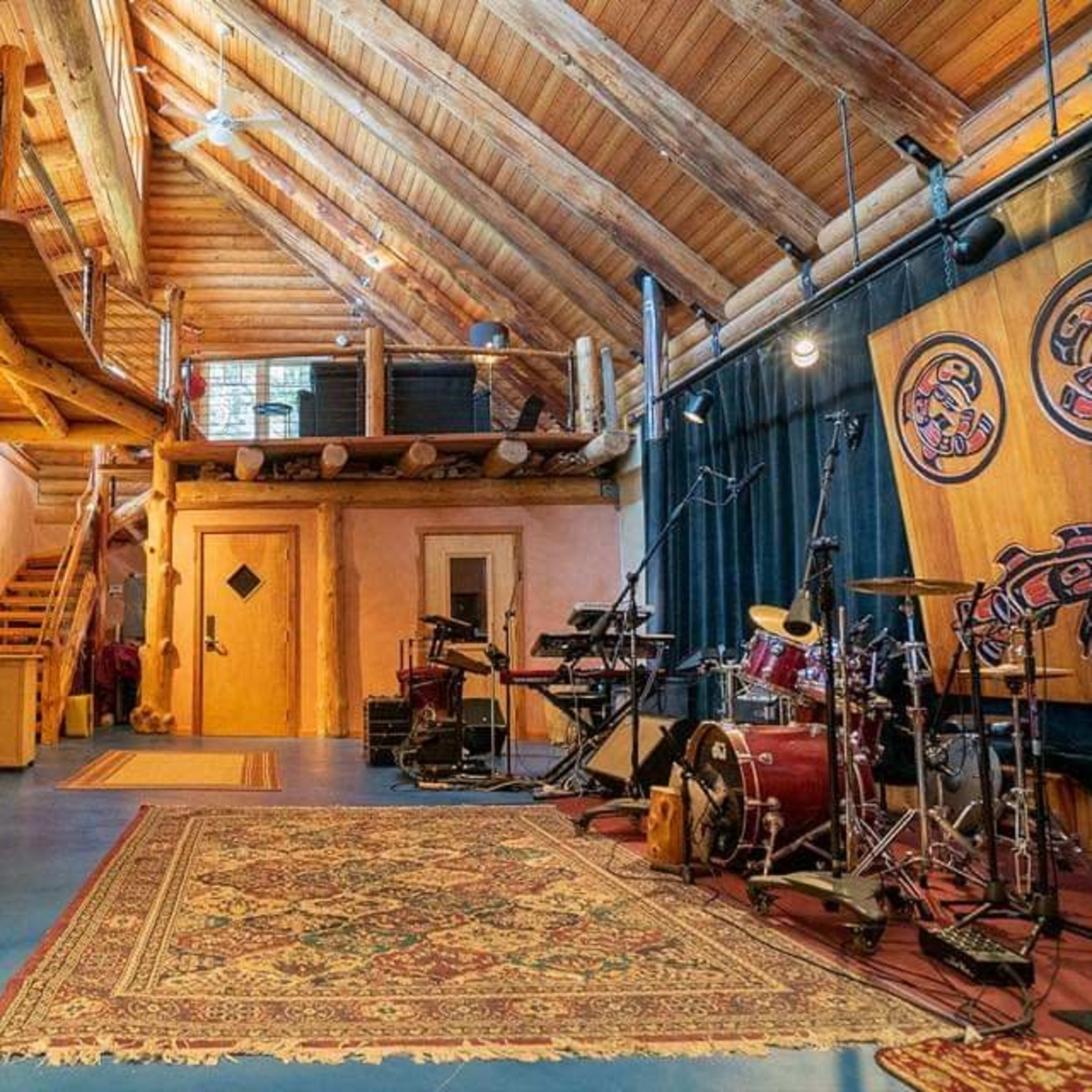 A spacious, wooden interior features a stage with musical equipment, a patterned rug, and a staircase leading to an upper level.