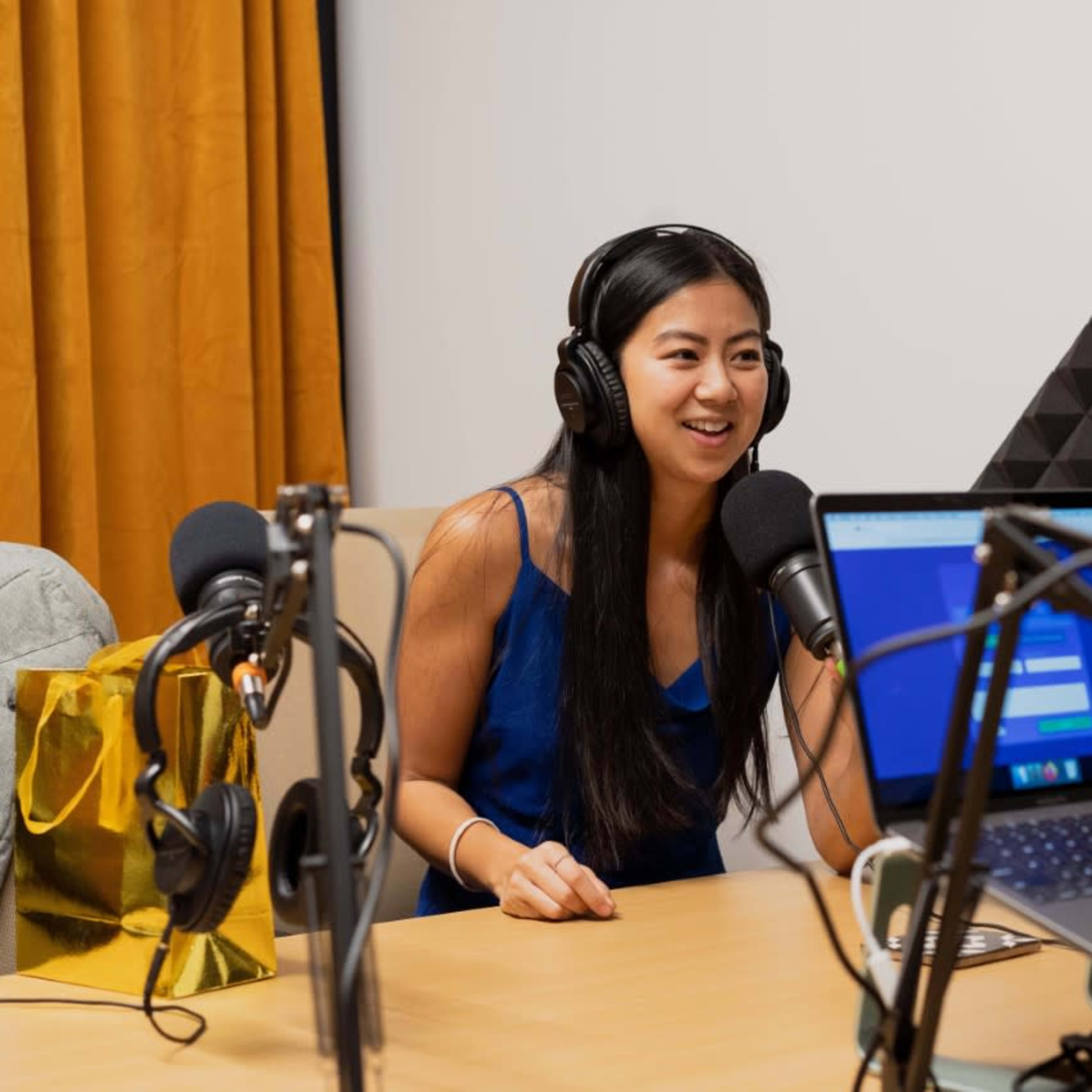 A woman wearing headphones sits at a table with microphones and a laptop in a podcast recording studio.