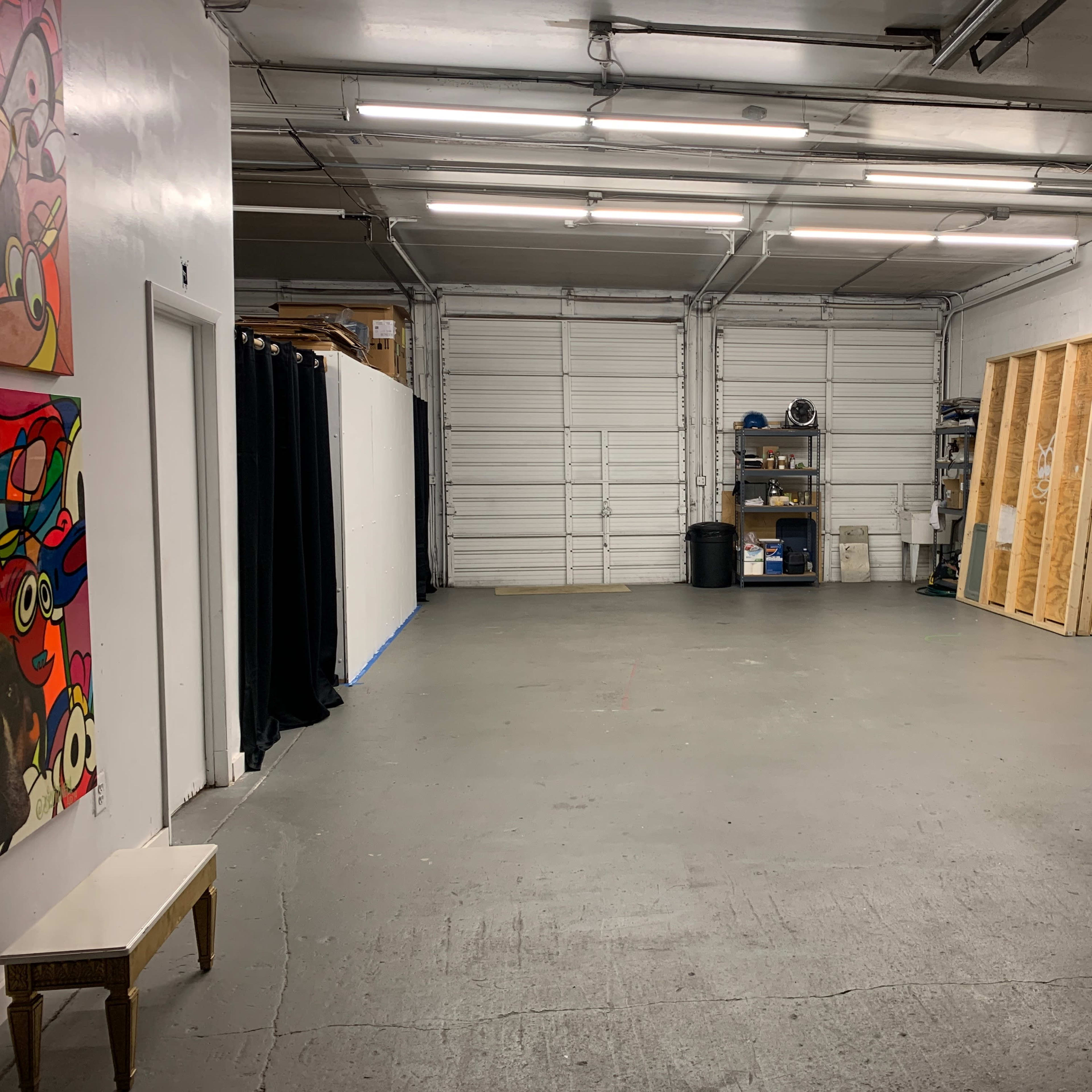 A spacious, empty room with concrete flooring, white walls, overhead lighting, and a mix of storage shelves and equipment along one side.