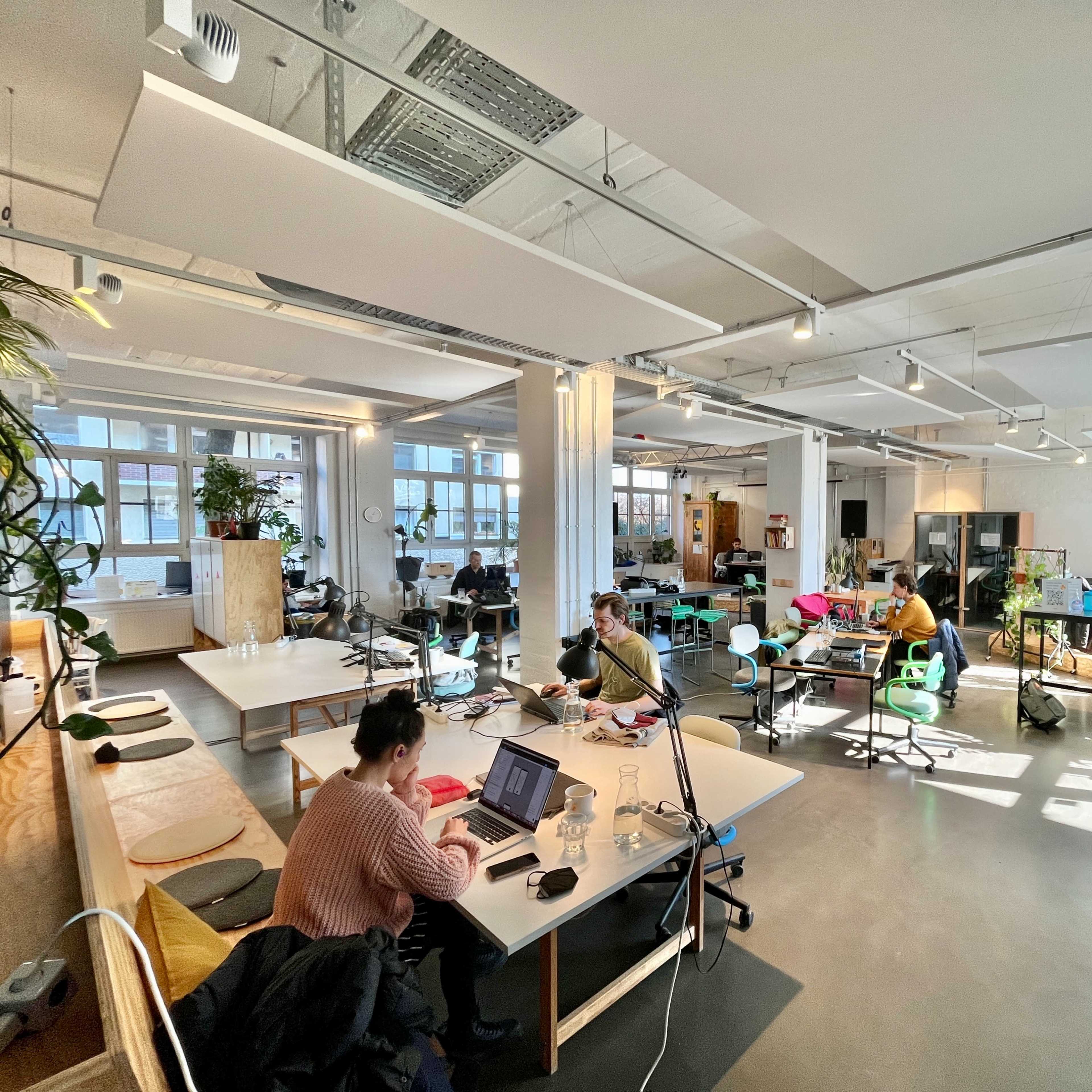A spacious office environment with multiple workstations, natural light streaming in through large windows, and several people engaged in various tasks.