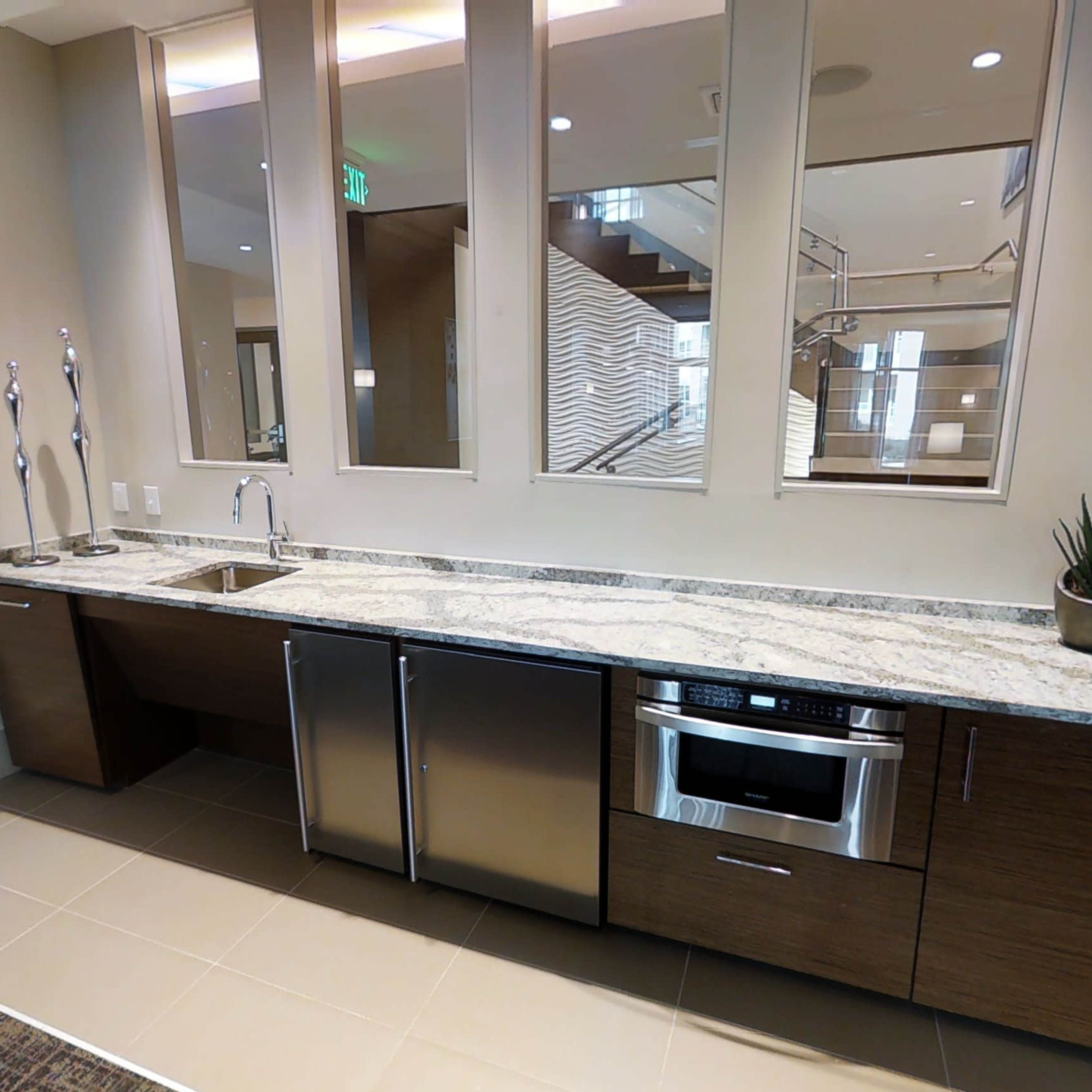 A modern kitchen area featuring a long countertop with a sink, stainless steel appliances, and a decorative plant, all set against a neutral wall with large windows.