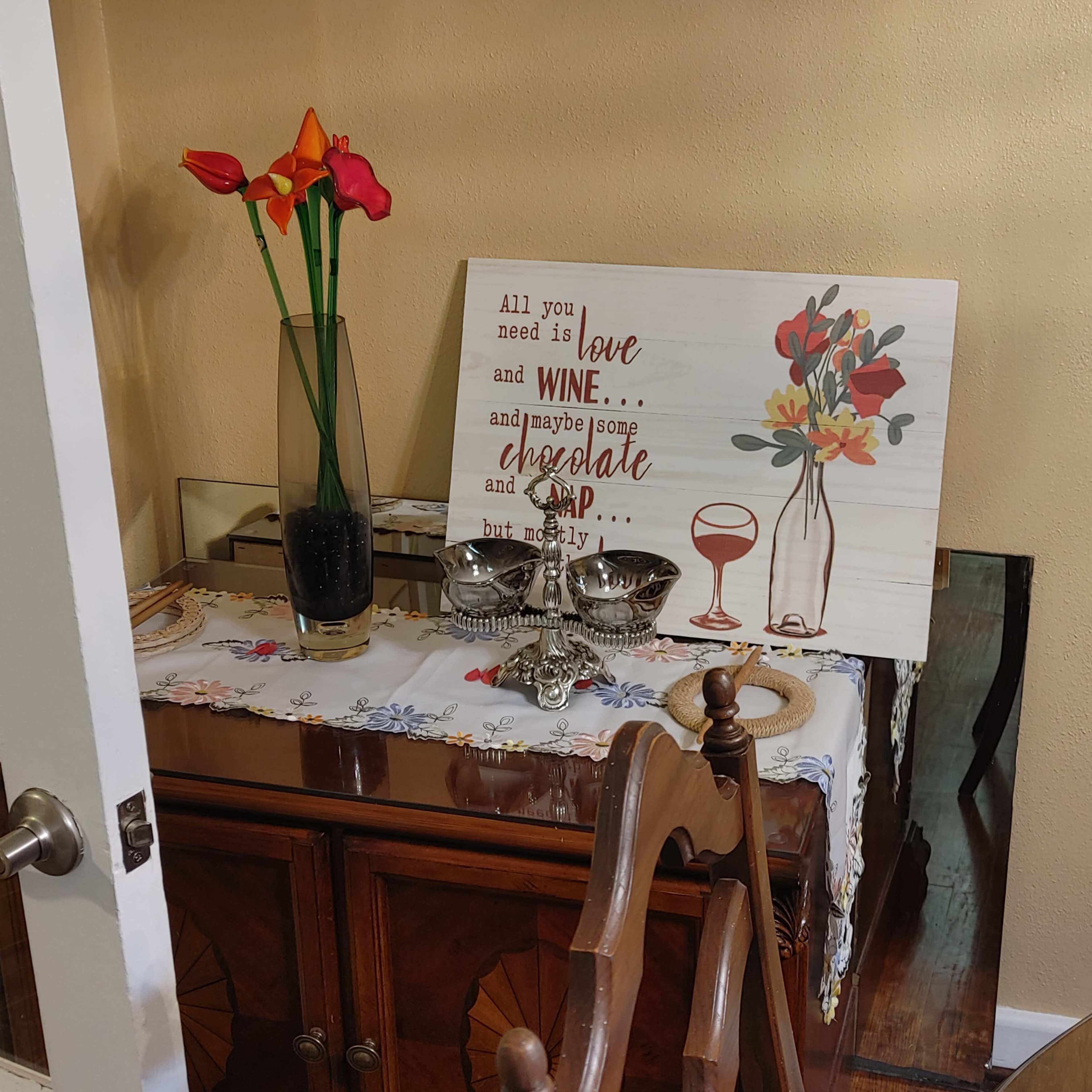 A sideboard features a vase of flowers, a decorative sign about love and wine, and several silver dishes on a lace tablecloth.