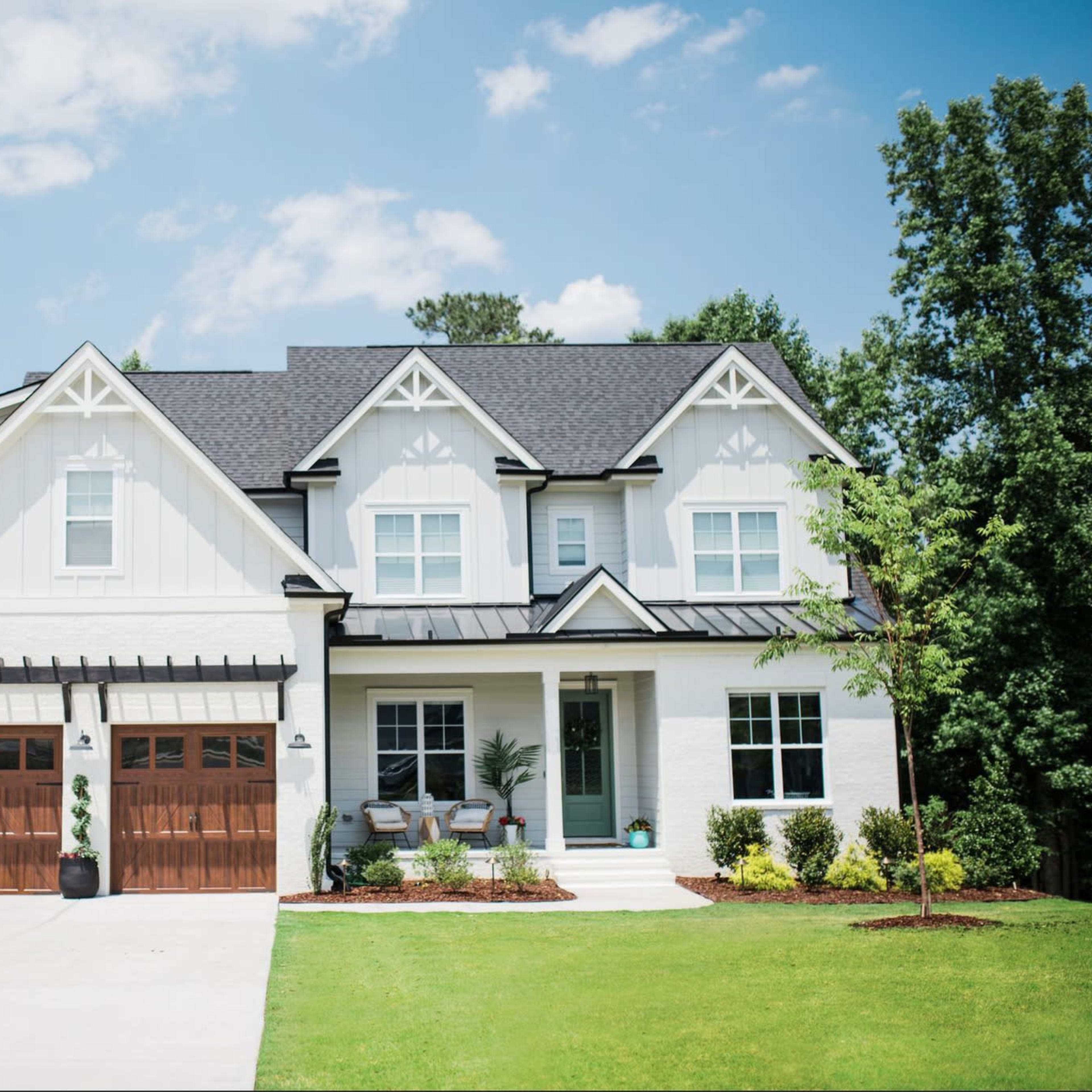A two-story white house with a black roof and wooden garage doors, surrounded by a well-manicured lawn and trees.