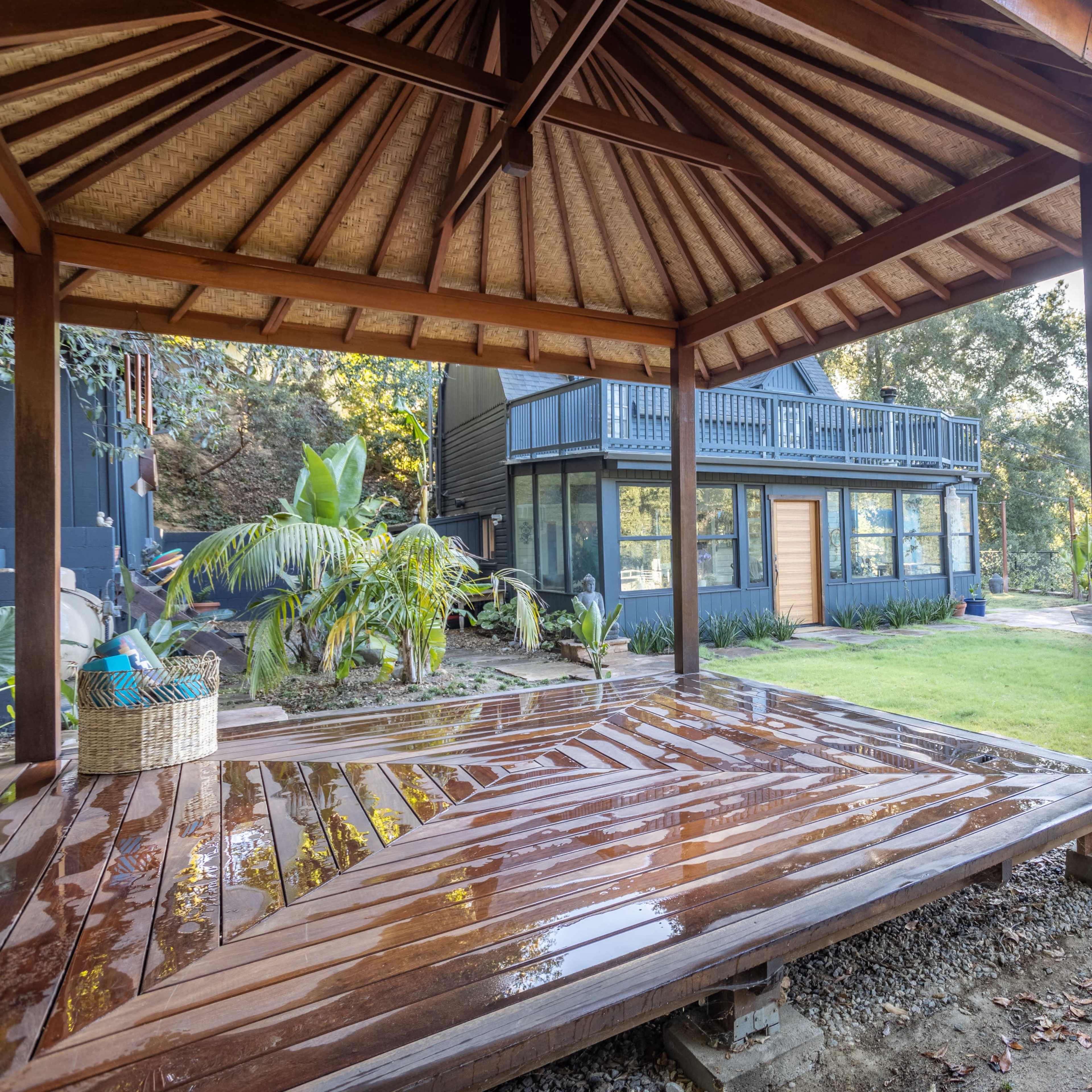 A wooden gazebo with a thatched roof overlooks a landscaped garden and a two-story house in a hilly area.