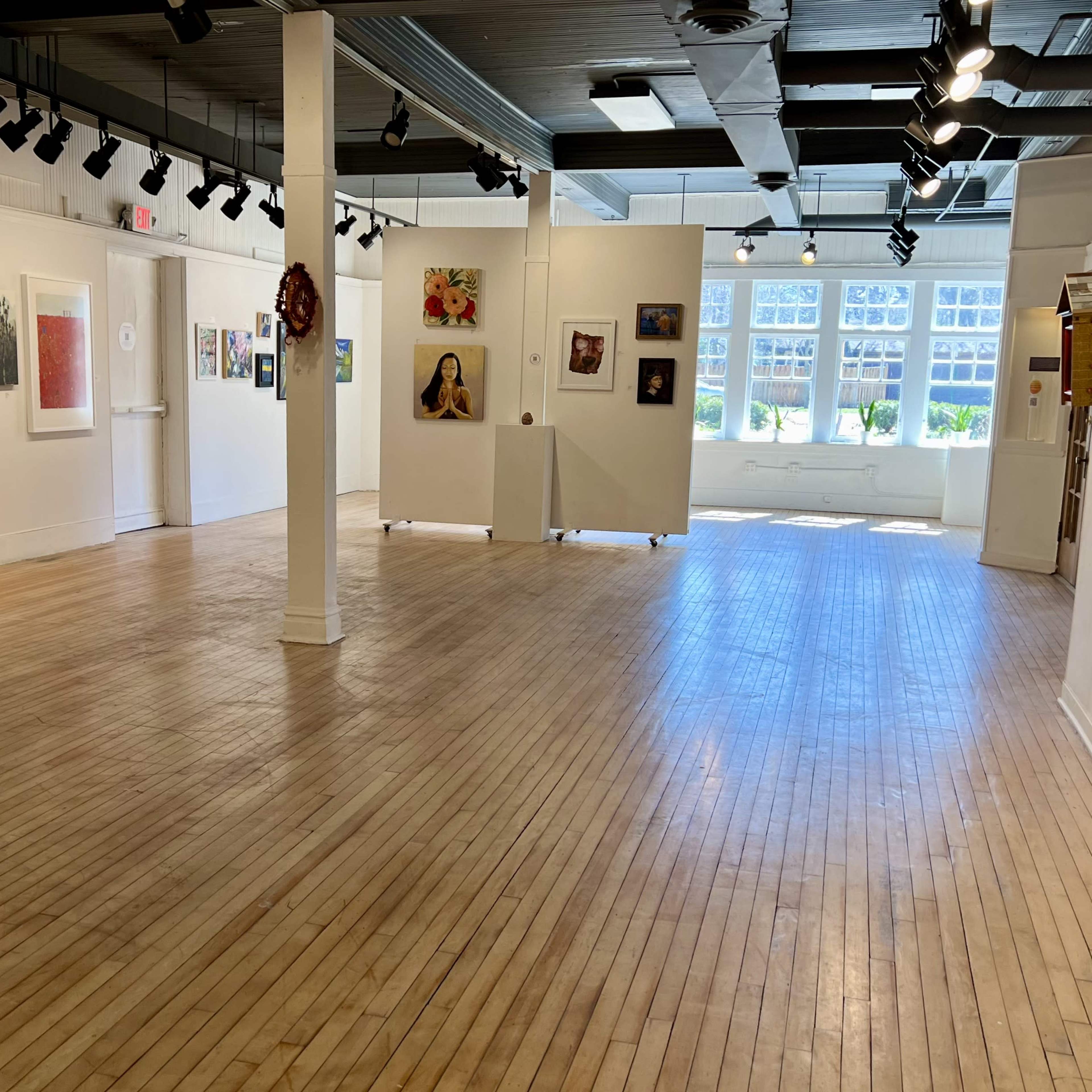 A spacious art gallery with wooden floors and white walls displaying various framed artworks, illuminated by overhead lighting.