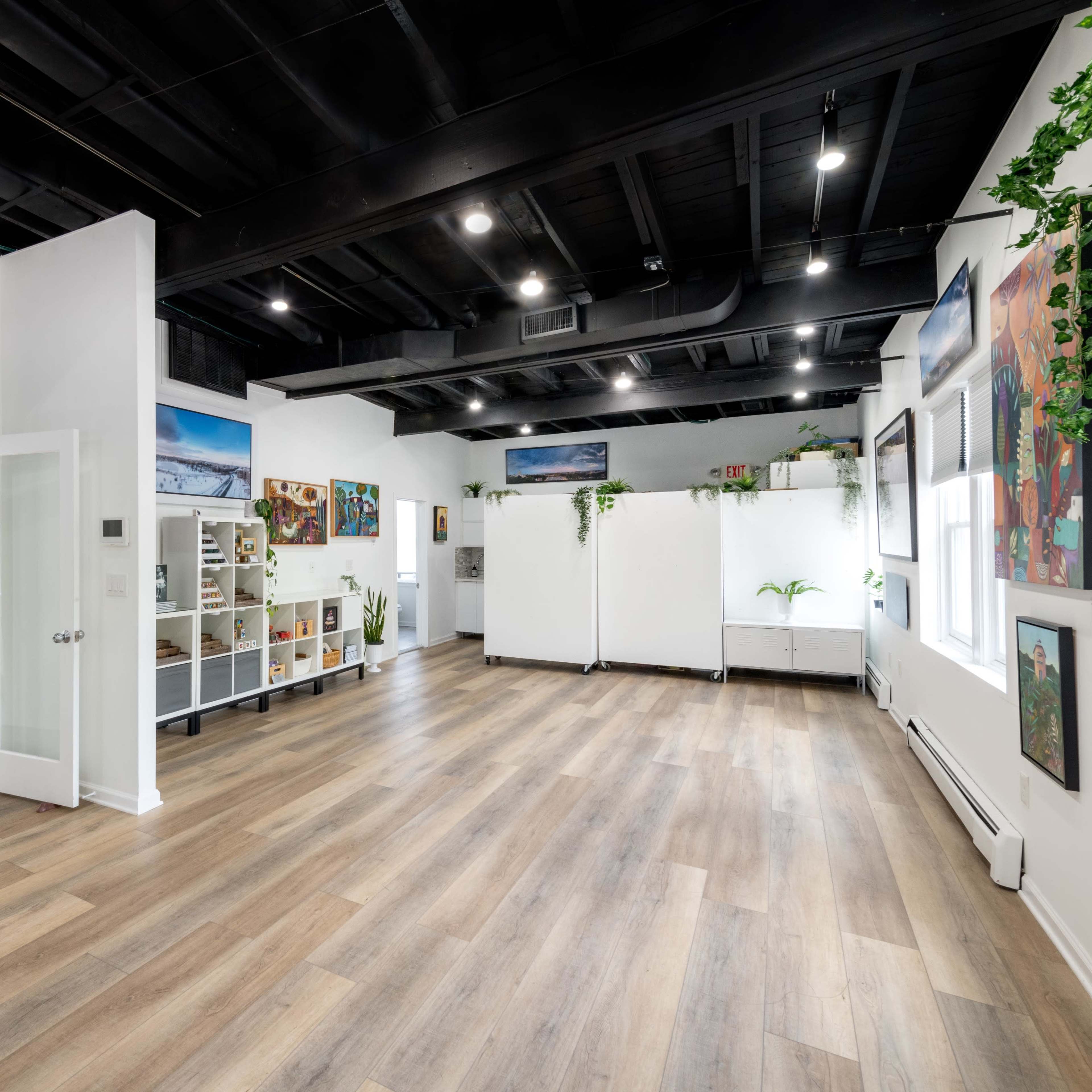 A spacious, well-lit room with wooden flooring, white walls, and plants, featuring artwork on the walls and a shelf along one side.