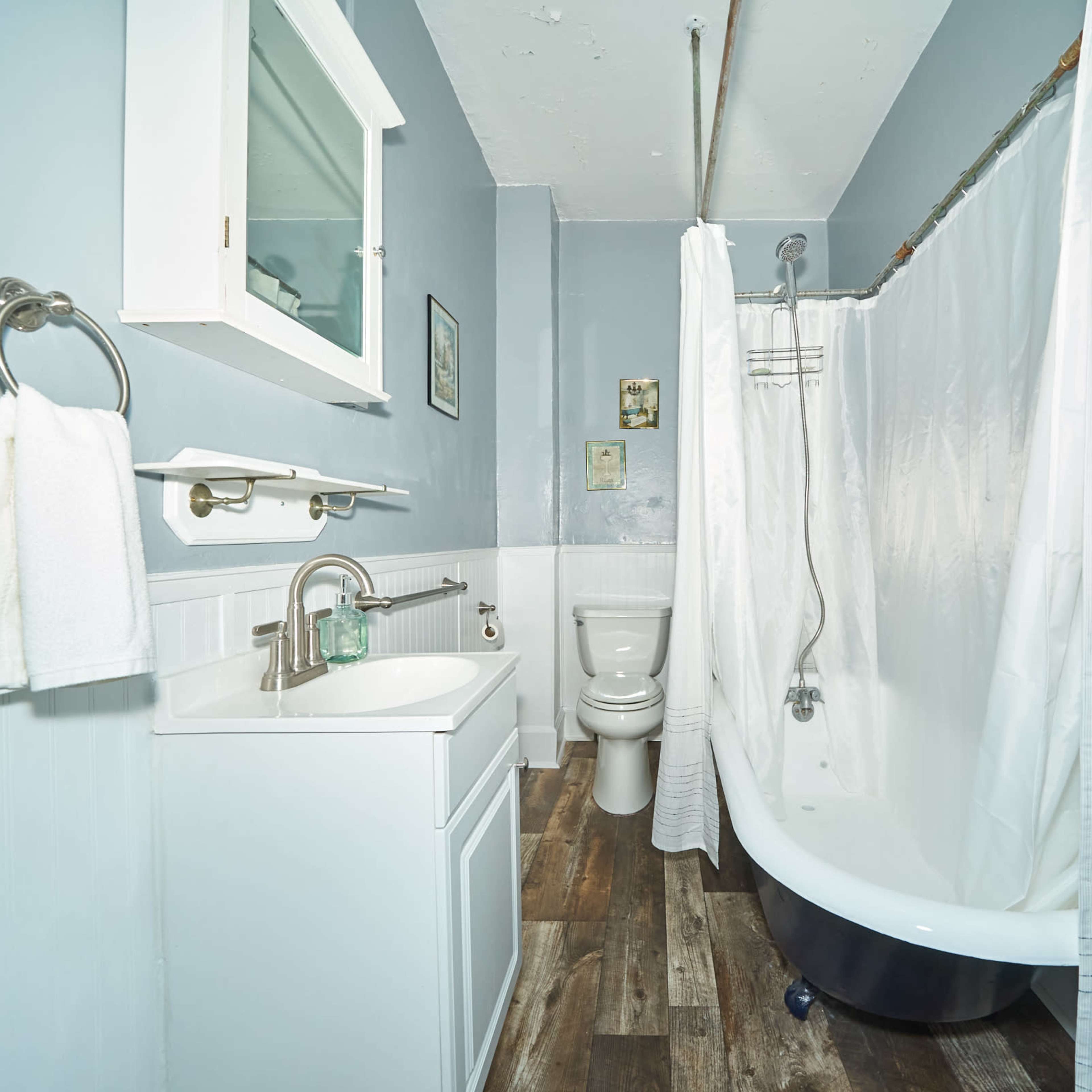A small bathroom featuring a shower with a white curtain, a toilet, and a sink with a cabinet, all set against light blue walls.