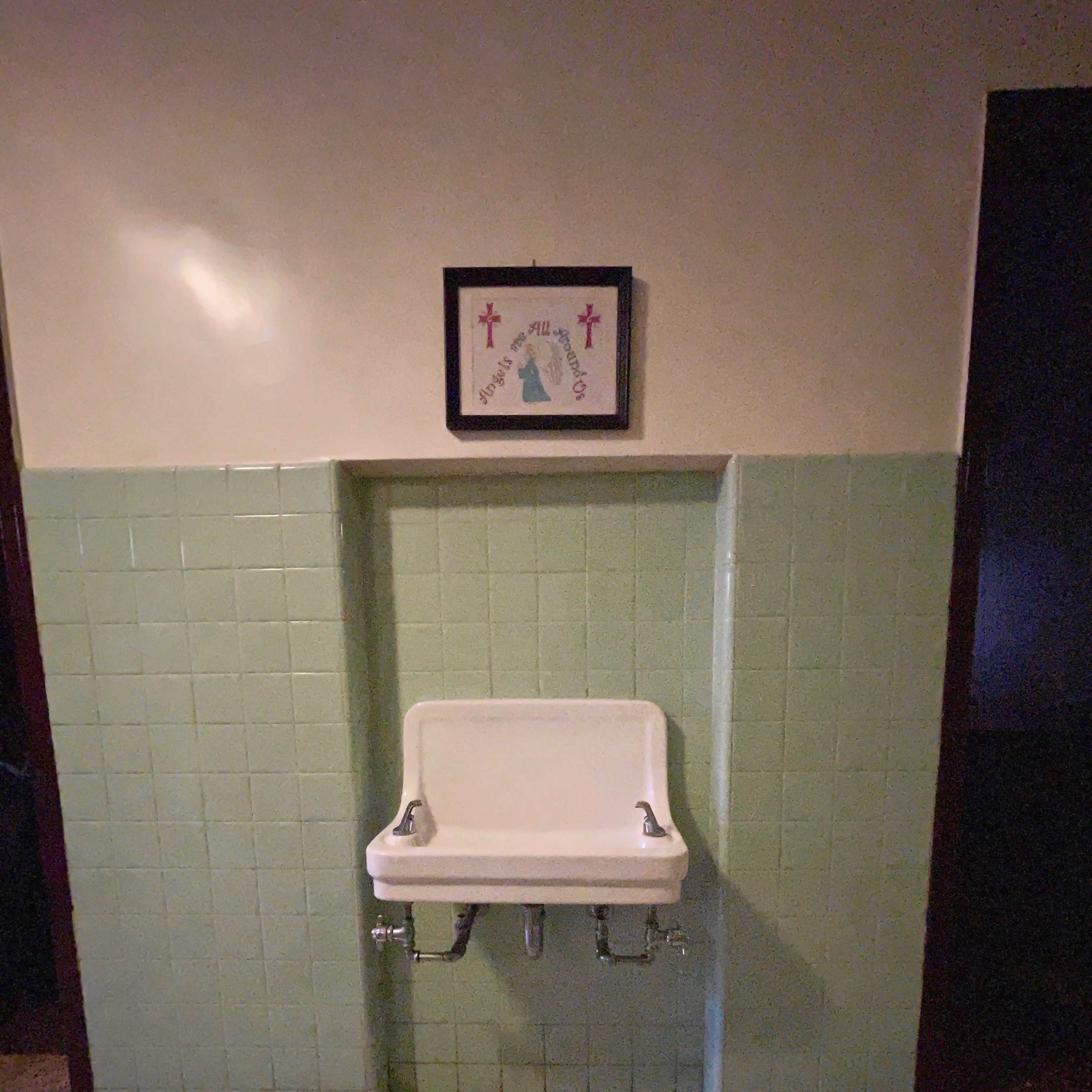 A white sink mounted on a pale green tiled wall beneath a framed decorative piece.