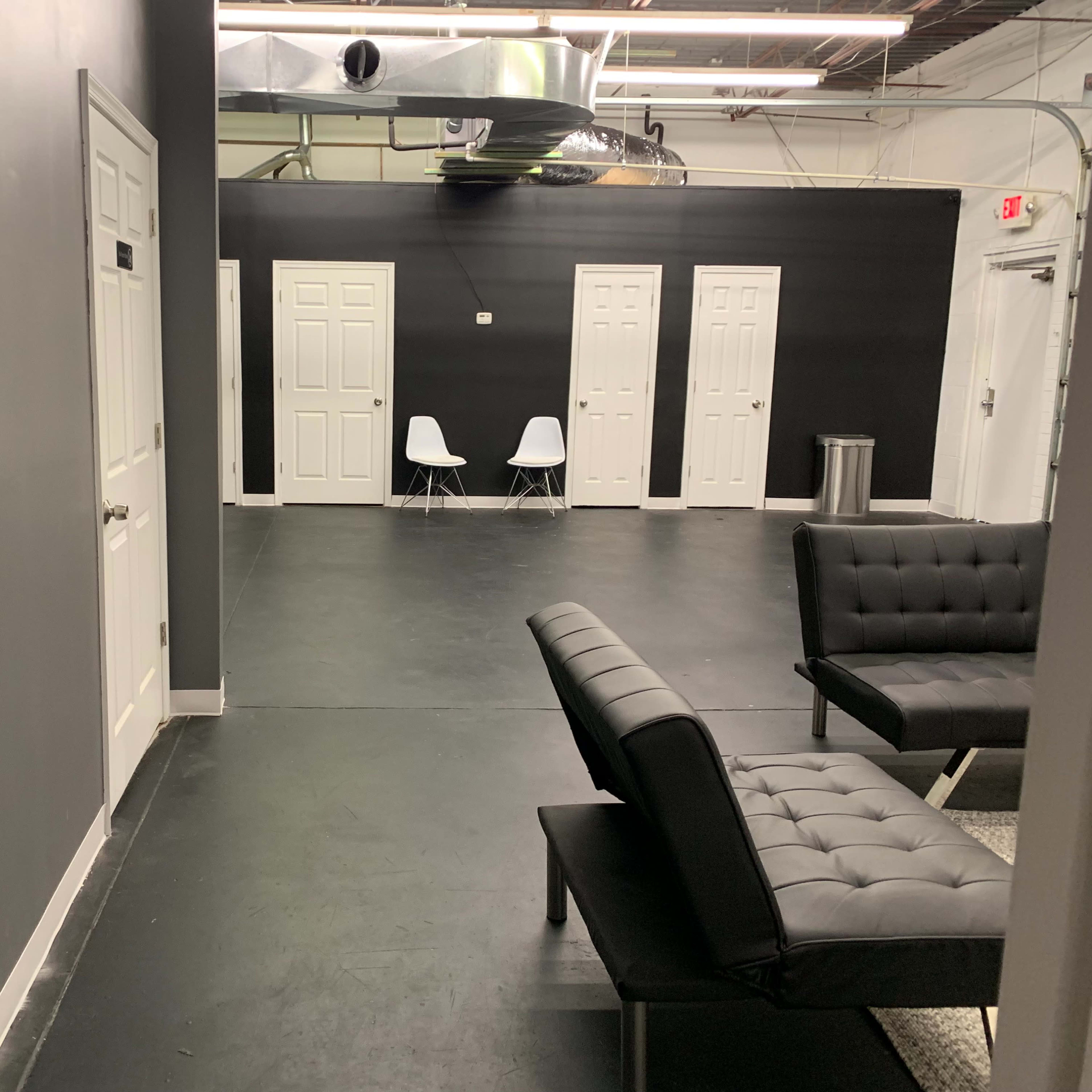 A corridor with gray walls, several closed doors, two white chairs, and black seating along the sides.