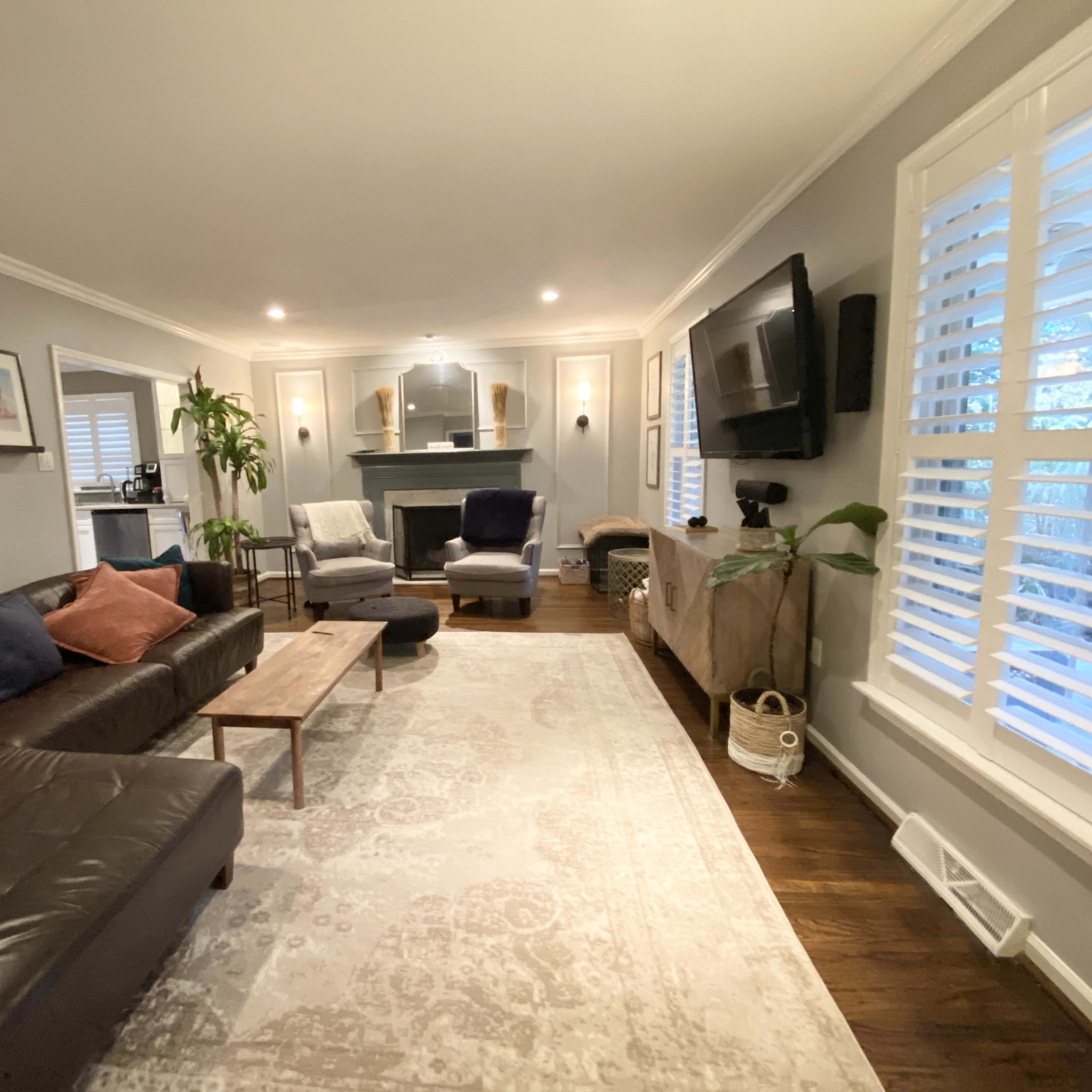 The living room features a dark leather sofa, a wooden coffee table, and a cozy seating area with two armchairs, framed artwork on the walls, and large windows with plantation shutters.