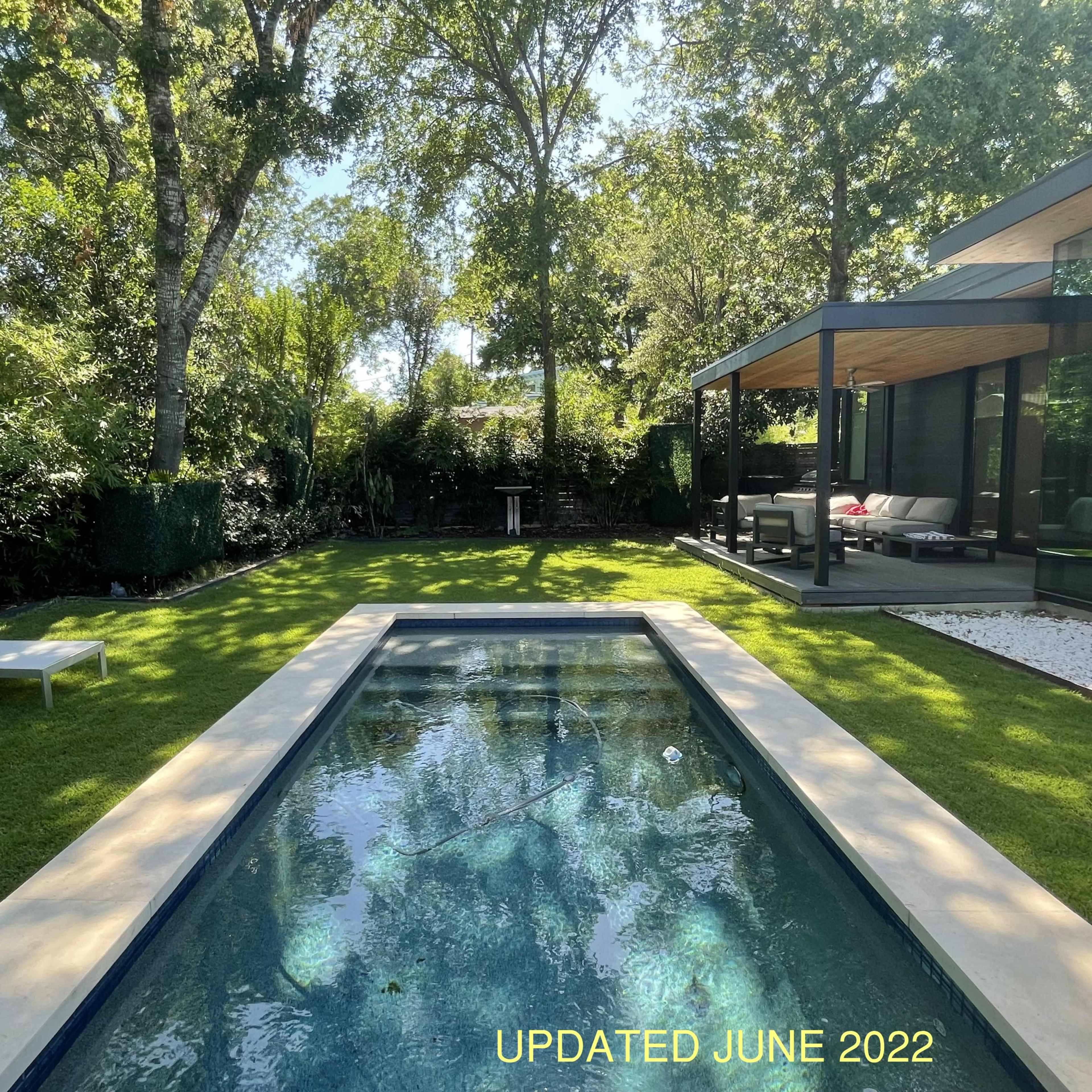 A modern backyard featuring a rectangular pool surrounded by neatly trimmed grass and lounge chairs, alongside a shaded seating area under trees.