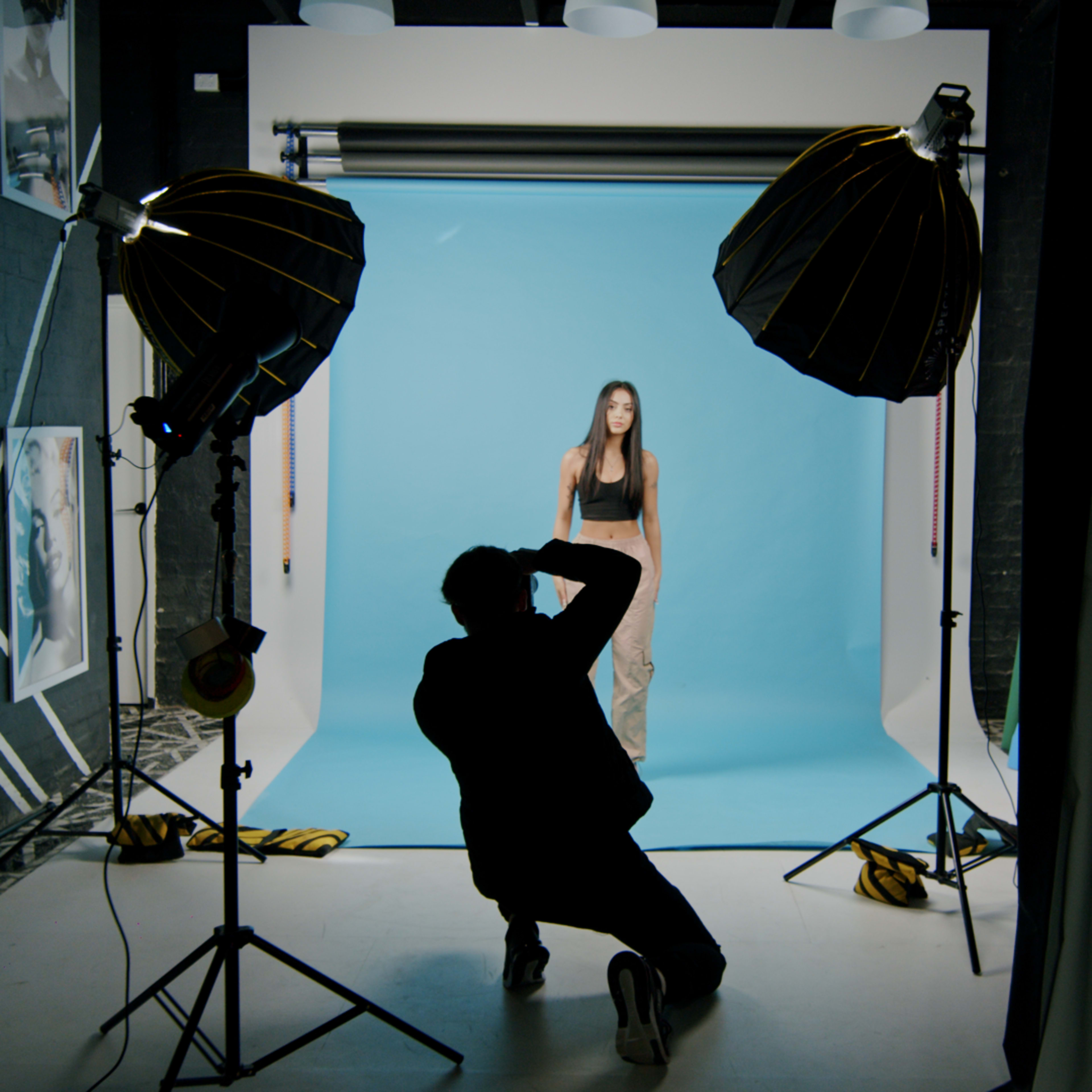 A photographer is capturing images of a model against a blue backdrop in a studio setup with lighting equipment.